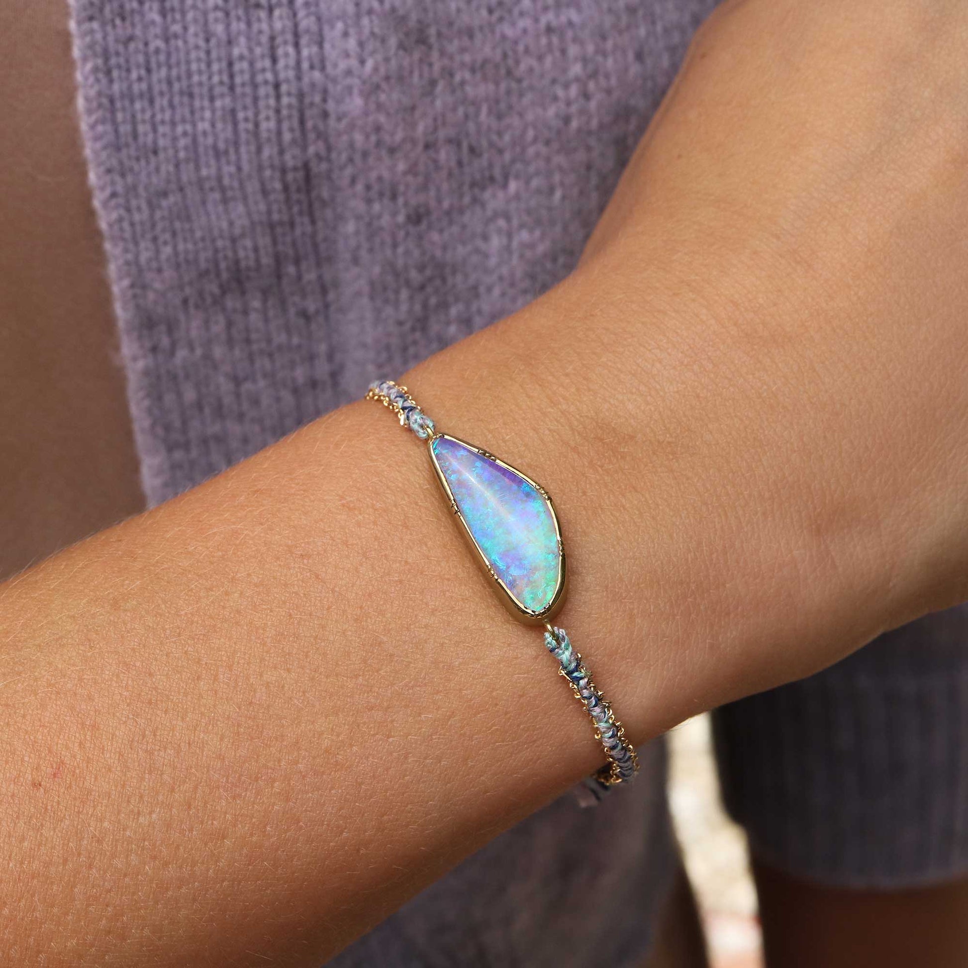 A delicate gold and silk bracelet with a ellipse-shaped iridescent boulder opal stone, shown worn on a person's wrist against a purple knit sweater.
