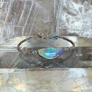 A handmade woven silk and 18k gold chain bracelet featuring a large, ellipse-shaped iridescent boulder opal stone in a gold bezel setting with a tassel button clasp, displayed on a clear crystal block.