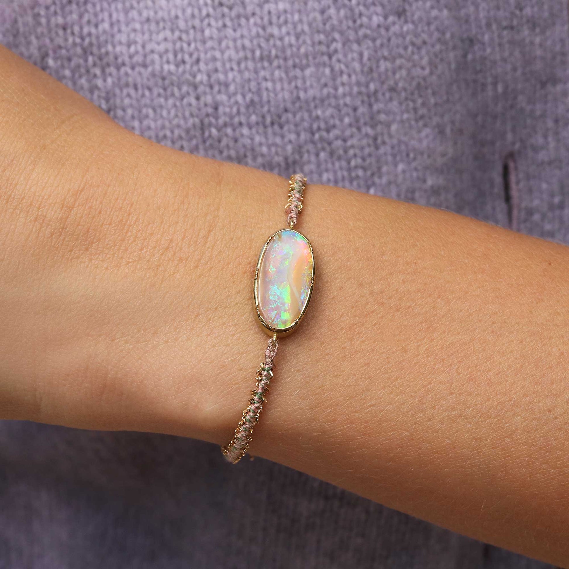 A delicate gold woven silk bracelet with a central iridescent opal stone, worn on a person's wrist against a textured gray knit background.
