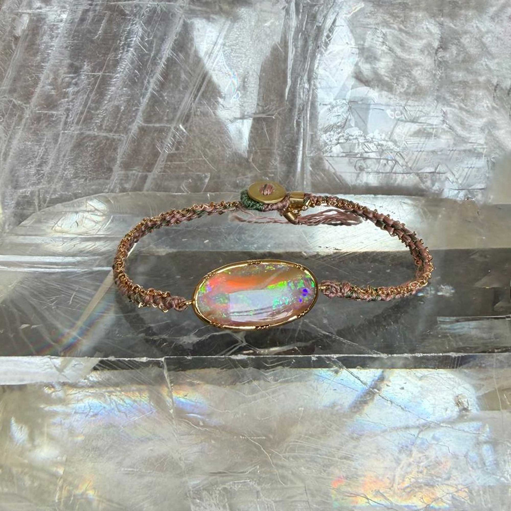A handmade woven silk bracelet with a large boulder opal centerpiece, resting on a reflective, iridescent surface.