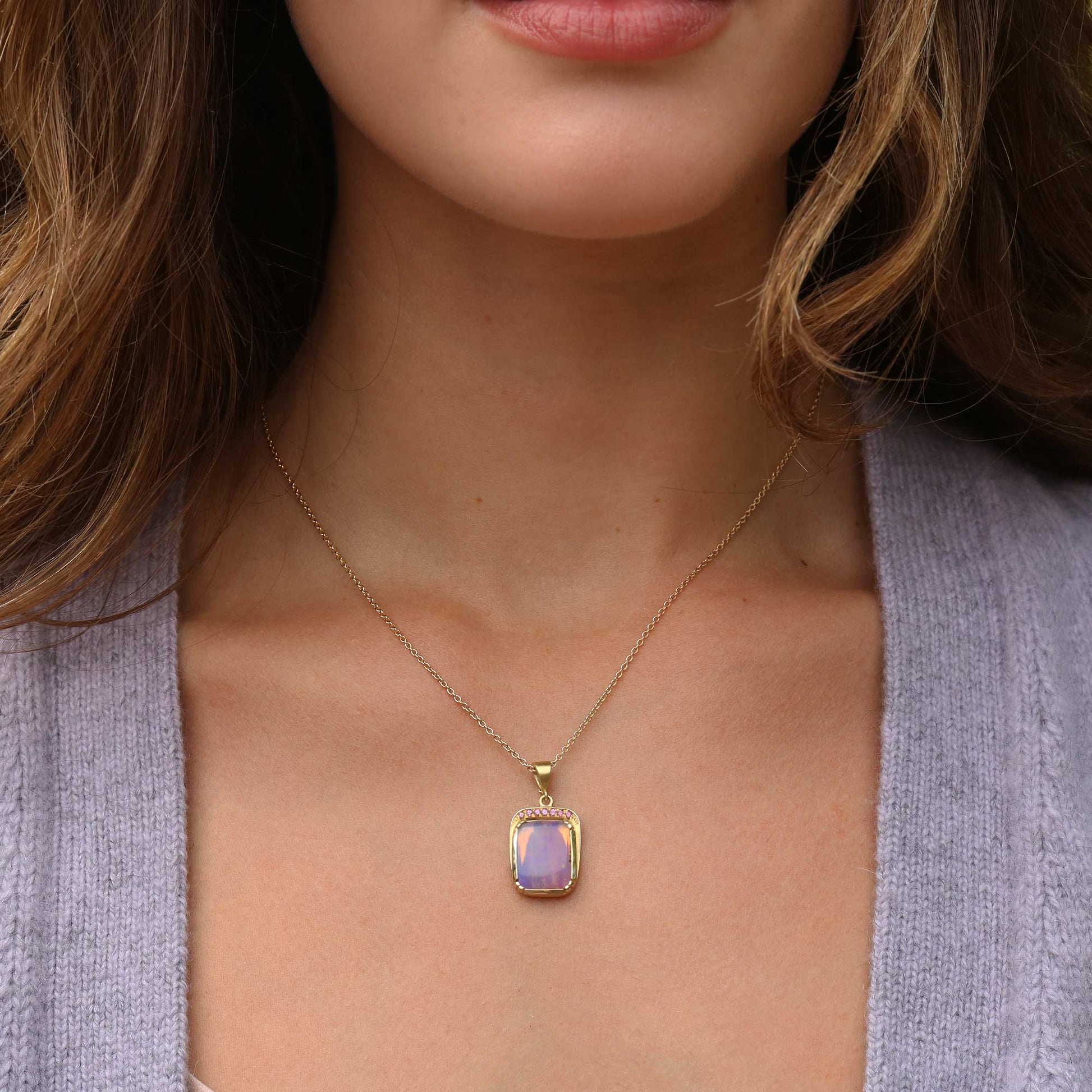 A close-up of a woman wearing a delicate gold chain necklace with a rectangular iridescent fossilised wood opal pendant that shifts between lavender and purple, set against her collarbone and a light purple cardigan.