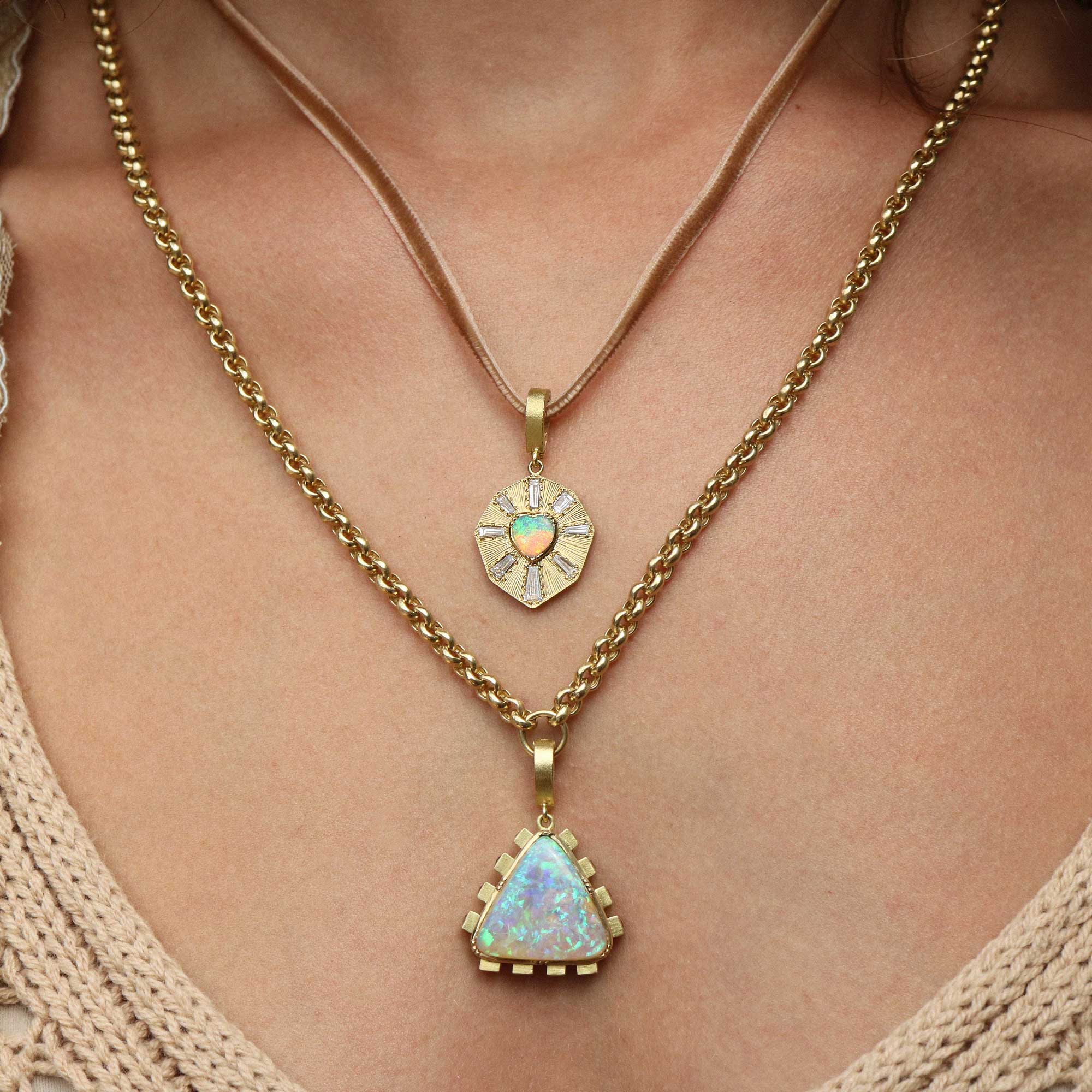 A close-up of a person's neck and chest wearing a layered gold necklace set. The shorter necklace has a small round pendant with a light blue opal-like center, while the longer necklace has a larger triangular pendant with a multicolored opal-like center.