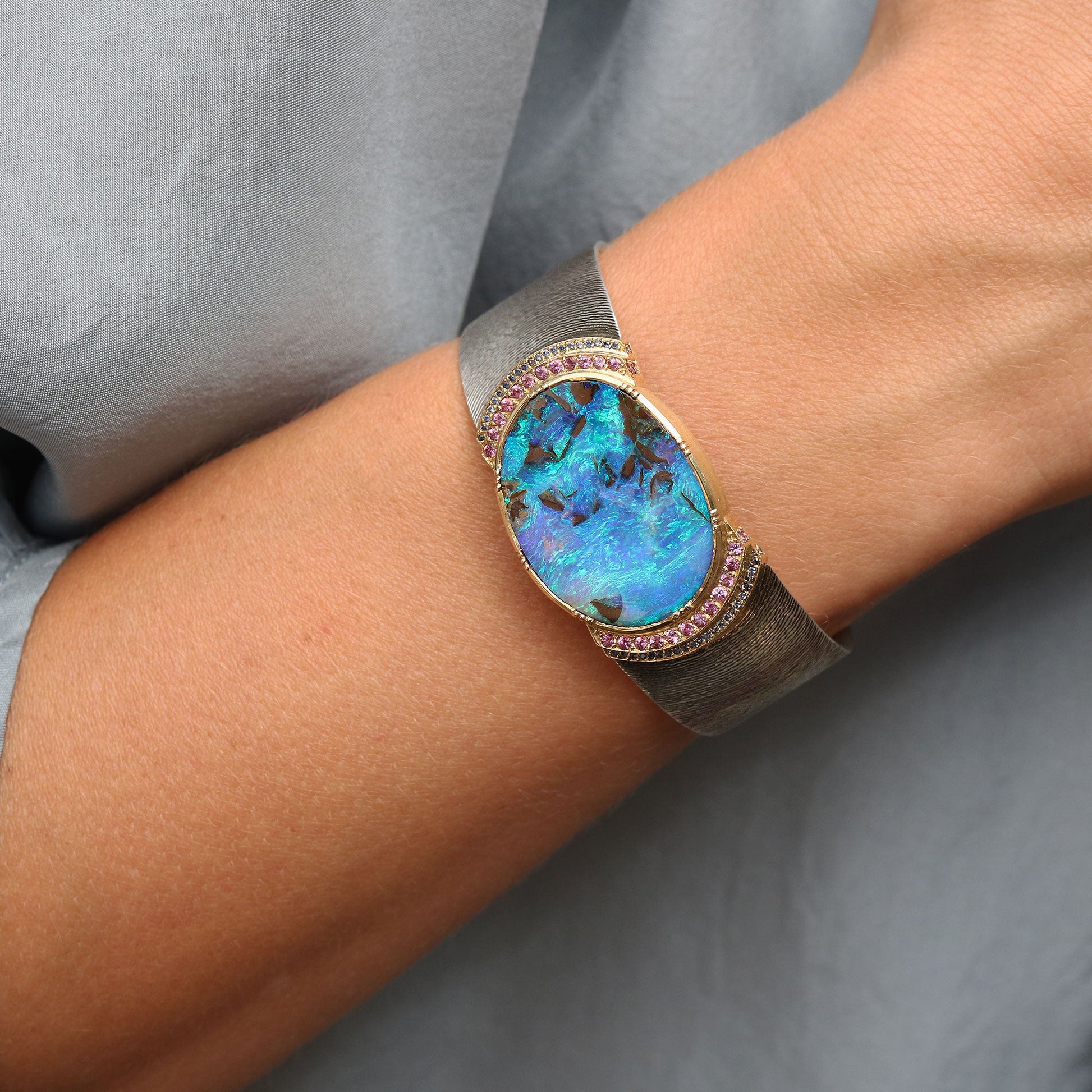 A close-up of a person's wrist wearing a wide silver cuff bracelet with a large oval blue opal center and pink gemstone accents around the bezel, against a soft gray fabric background.