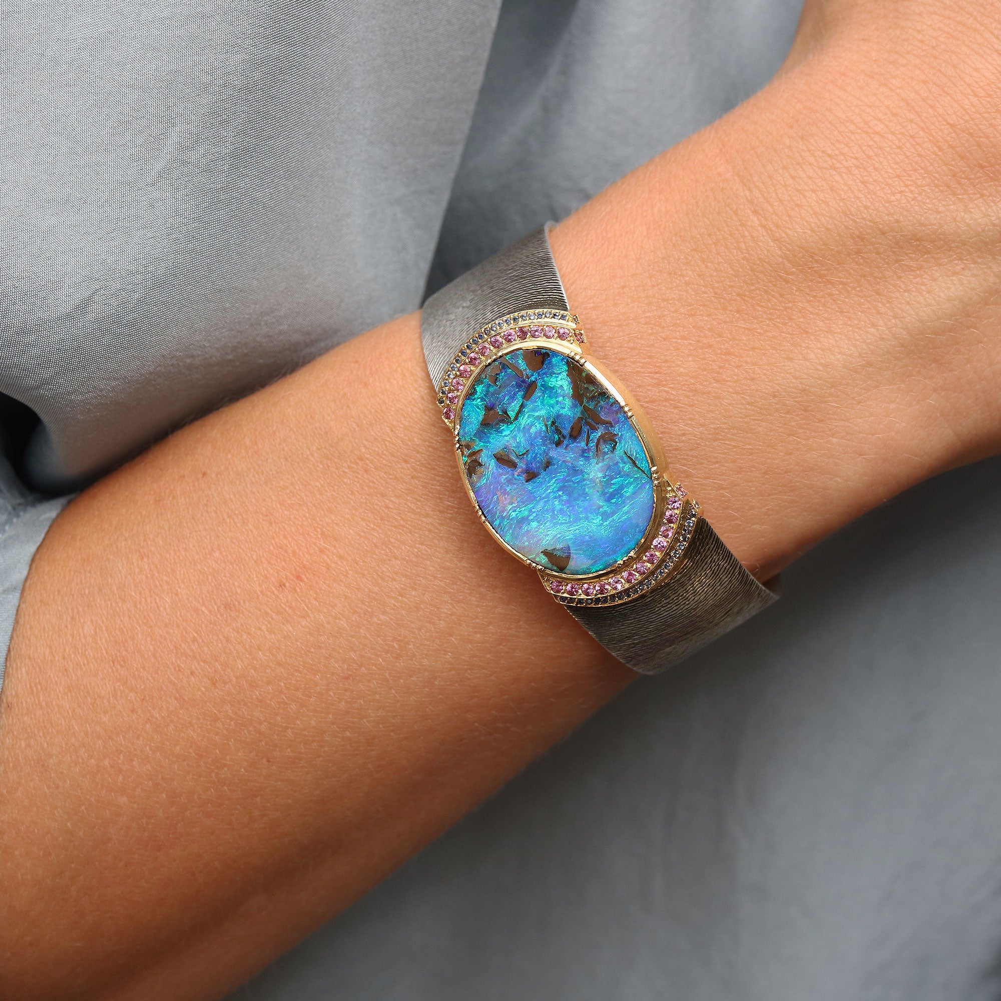 A close-up of a person's wrist wearing a wide silver cuff bracelet with a large oval blue opal center and pink gemstone accents around the bezel, against a soft gray fabric background.