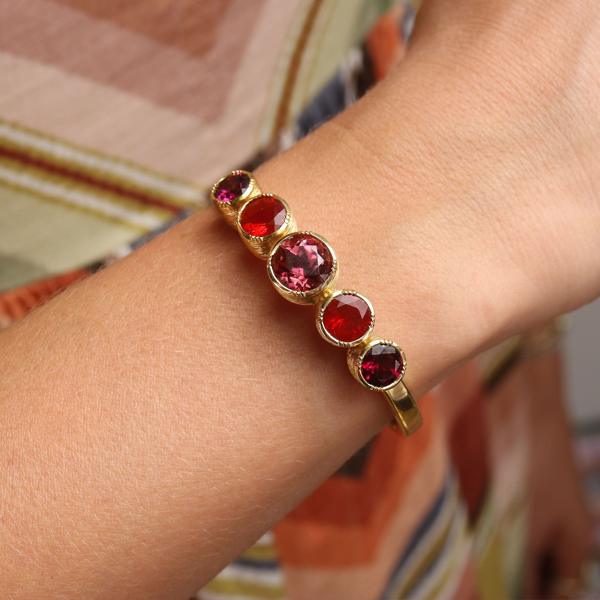 A gold bangle bracelet with four round red fire opal and tourmalines and garnet gemstones, worn on a person's wrist against a soft, blurred striped background.