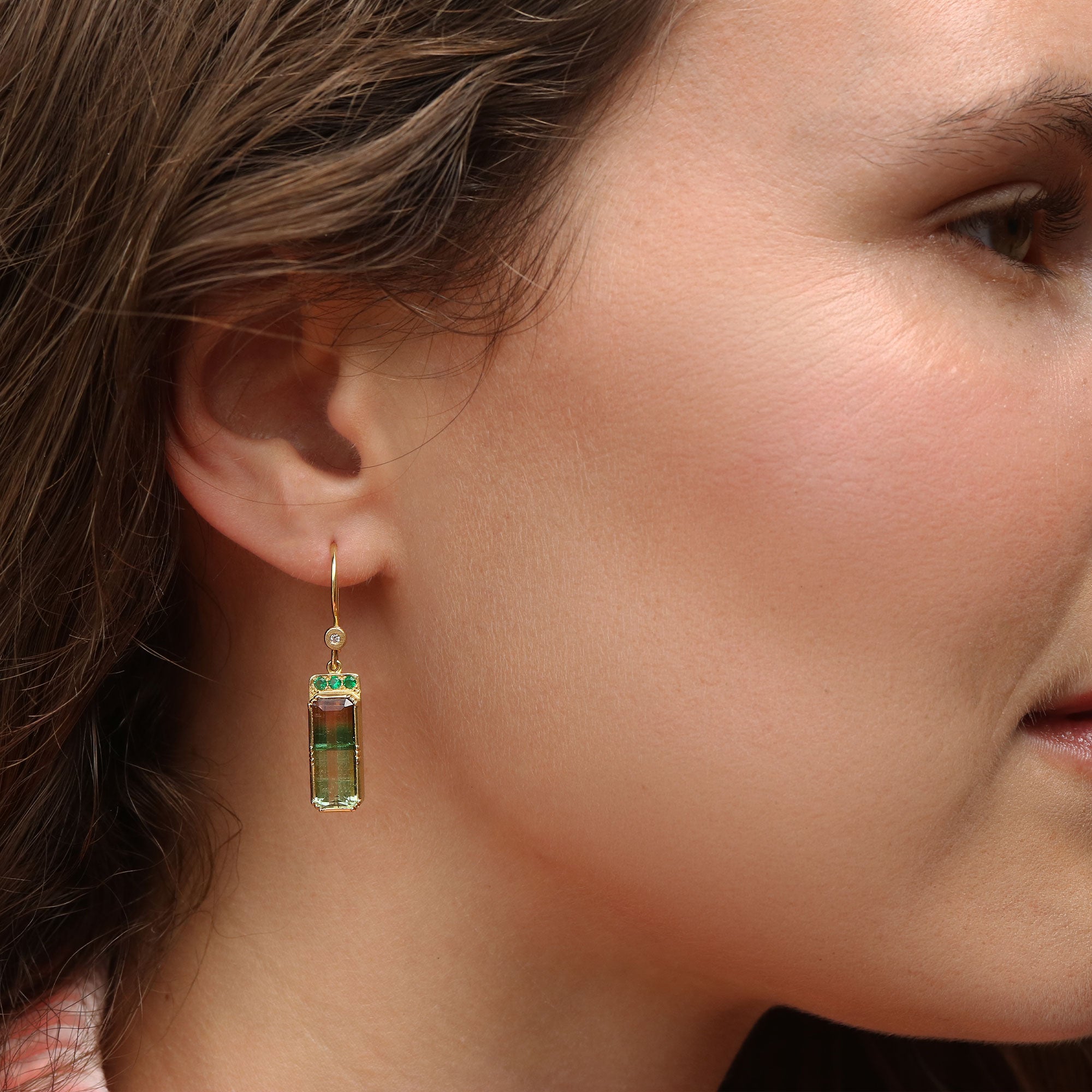 A close-up side profile of a woman wearing a single gold dangle earring with a bi colour tourmaline rectangular faceted gemstone with emerald accents and diamond above.