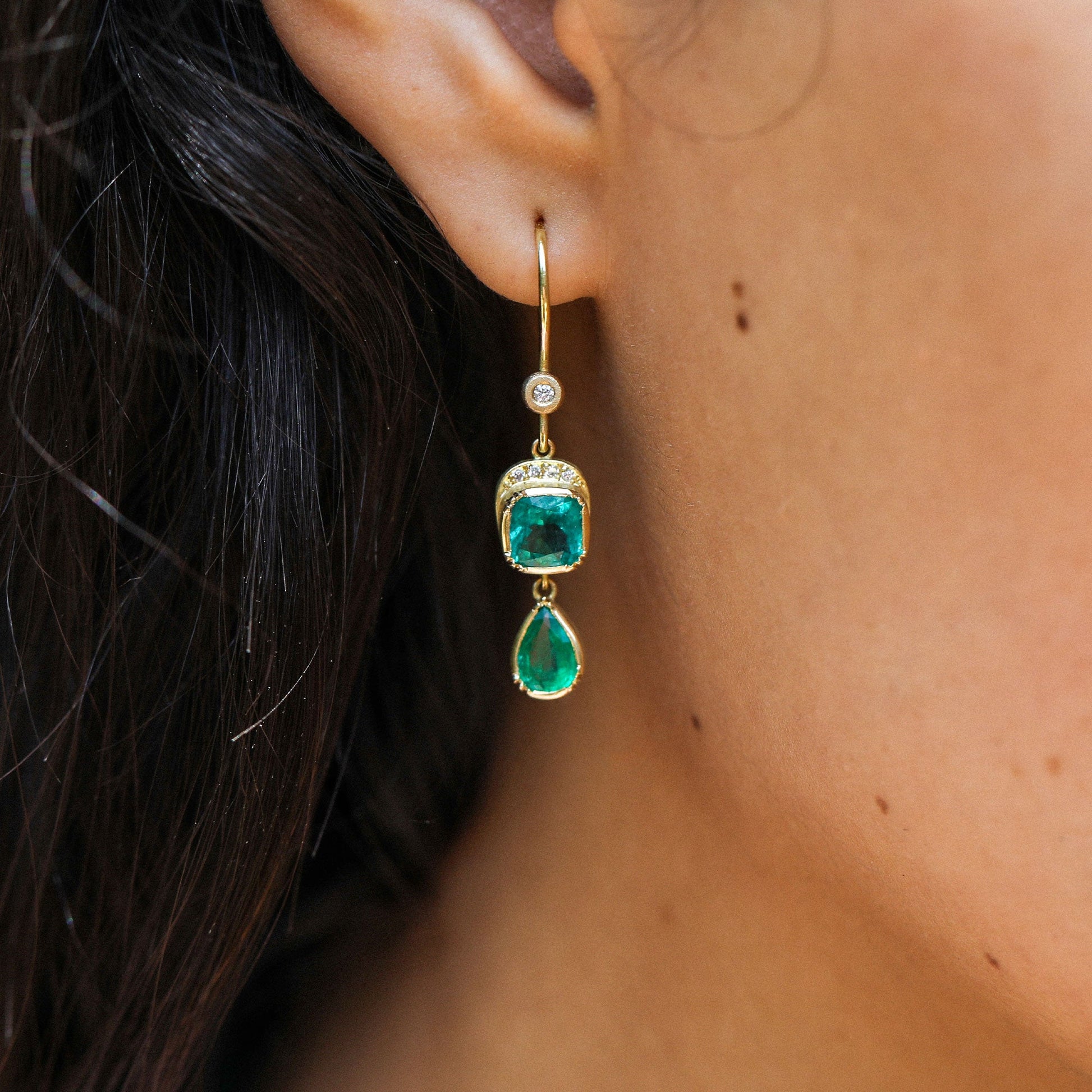 A close-up of a person's ear wearing a gold drop earring with a diamond stone and a green emerald-shaped drop with a teardrop-shaped green emerald stone below.