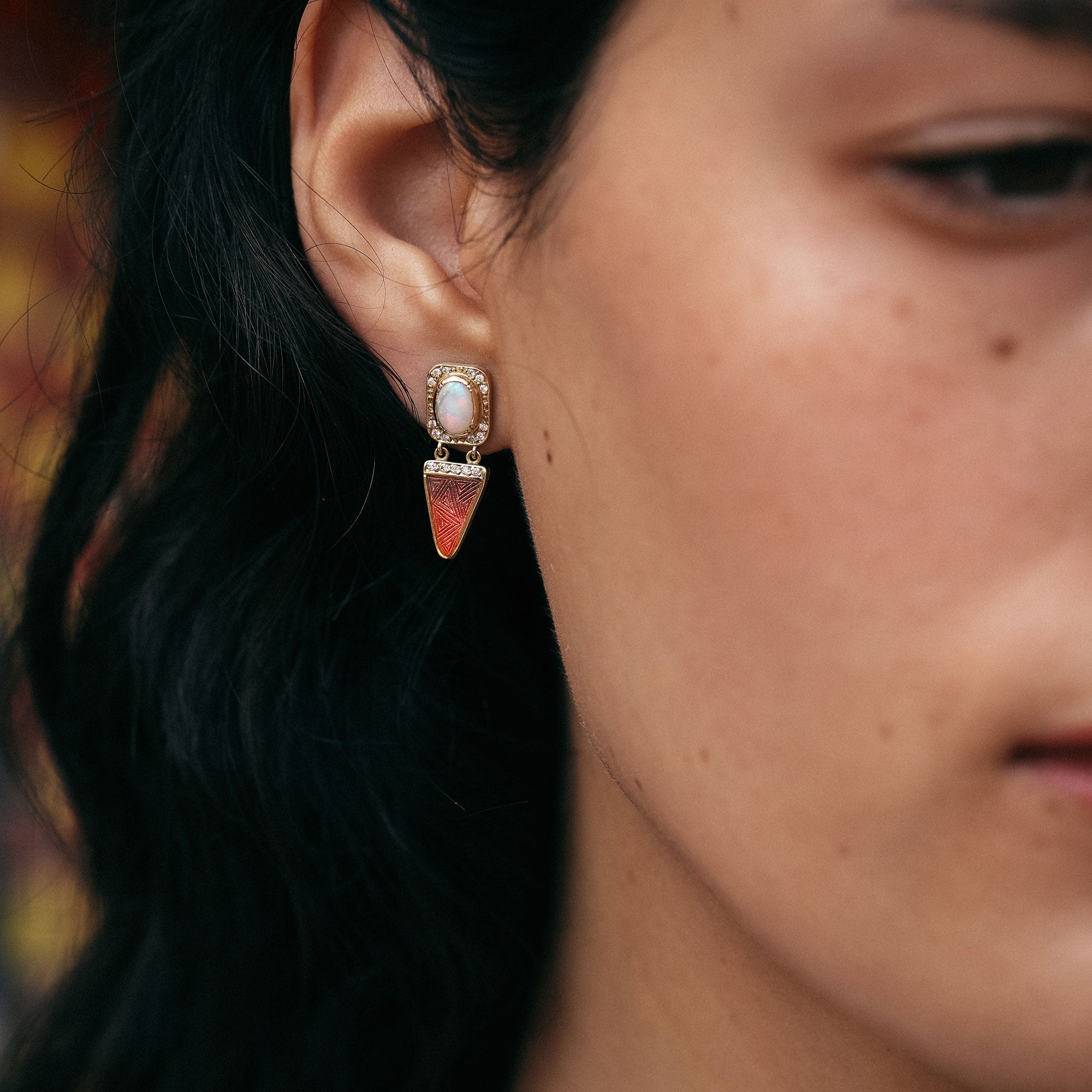 Close-up of a person wearing a decorative opal and enamel earring with a blurred background