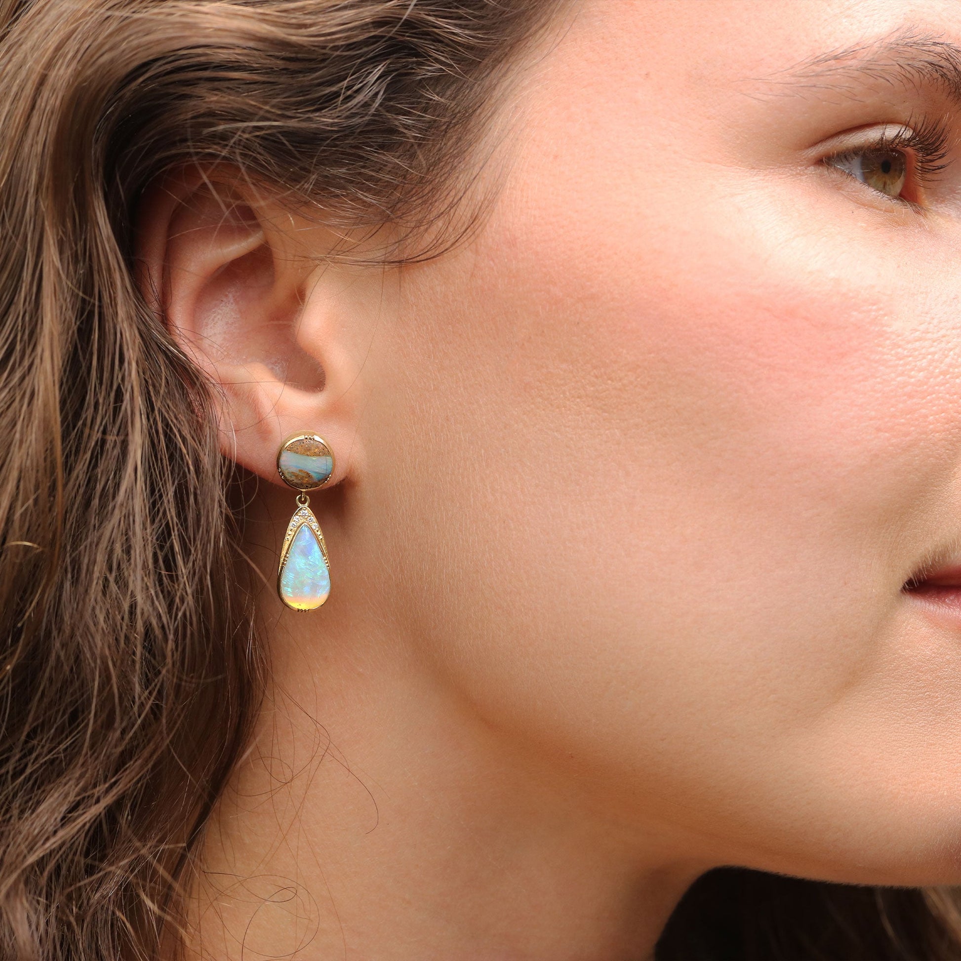 A close-up of a woman's ear wearing a gold drop earring with a round light blue opal top and a teardrop-shaped light blue opal drop.