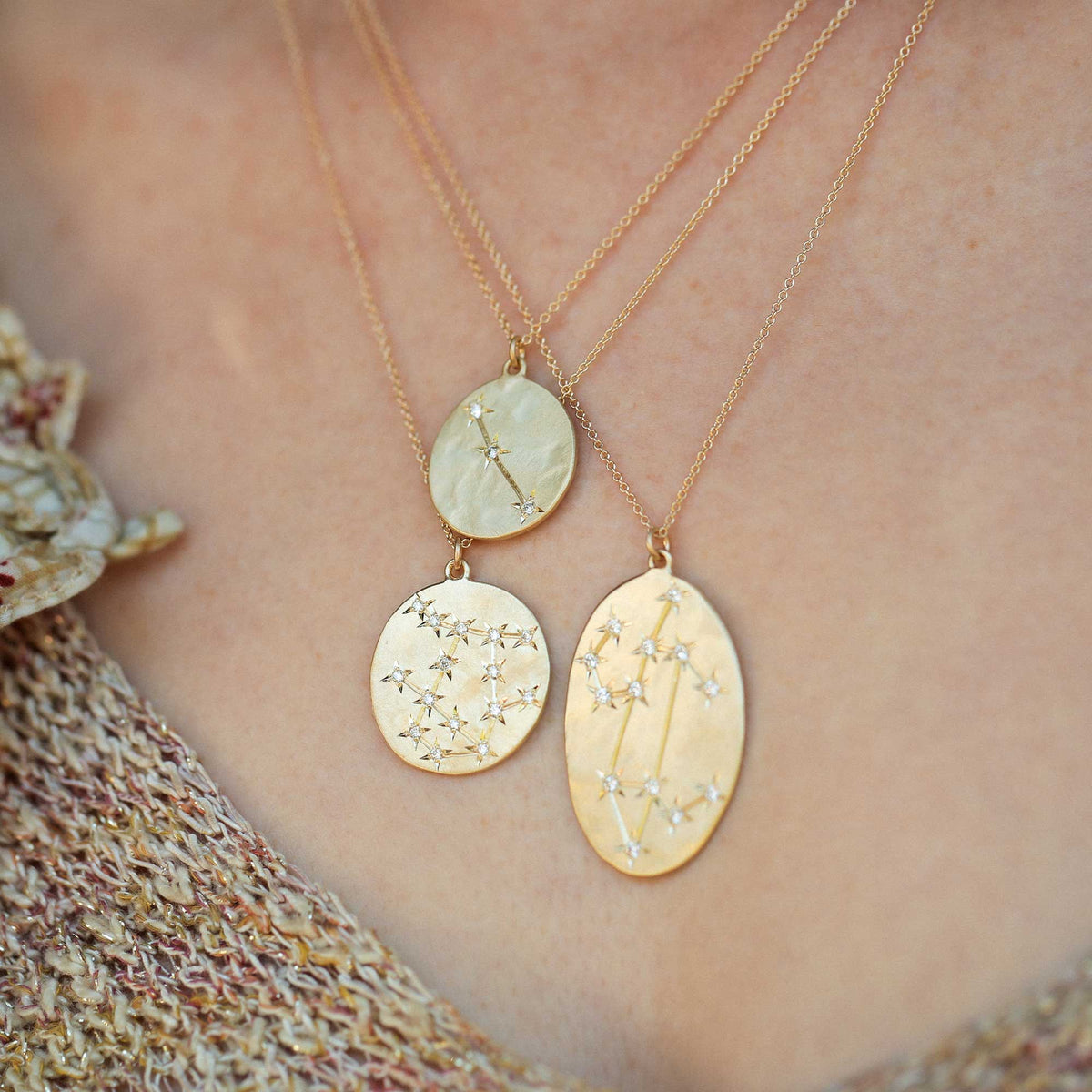 Three 14k gold oval pendant necklaces with engraved star and constellation designs, shown worn on a person's neck over a textured knit top.