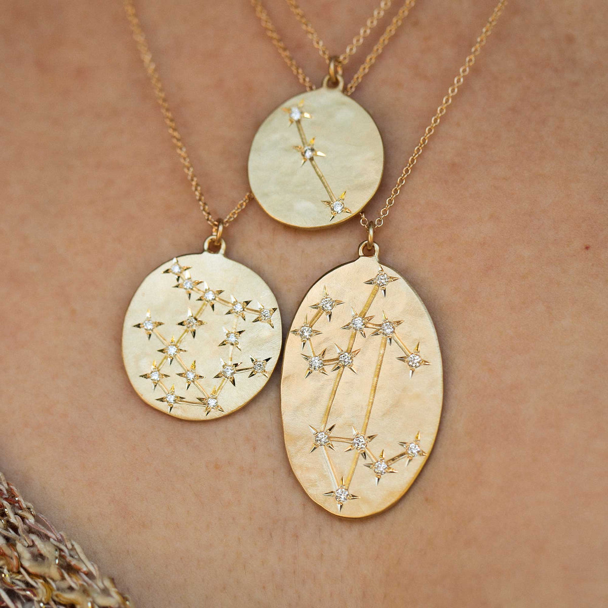 Three 14k gold constellations necklaces with textured oval pendants and small diamond stones, shown worn on a person's neck against a light skin tone background.