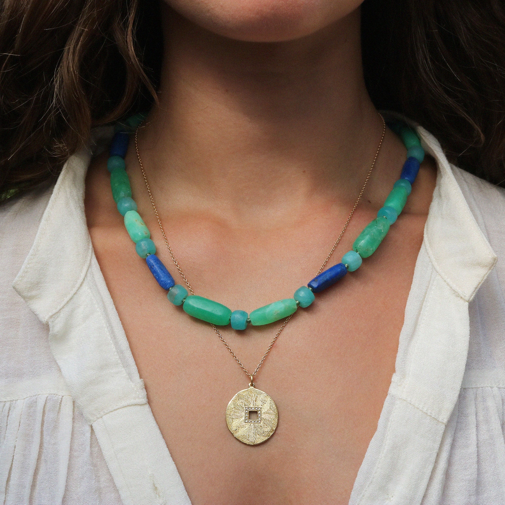 A close-up of a person wearing layered necklaces. The outer necklace features translucent green and blue beads, while the inner necklace is a delicate gold chain with a round gold coin pendant.