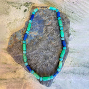 A beaded necklace made of alternating turquoise, light green, and royal blue cylindrical beads with a gold clasp, displayed on a textured gray stone against a light beige stone background.