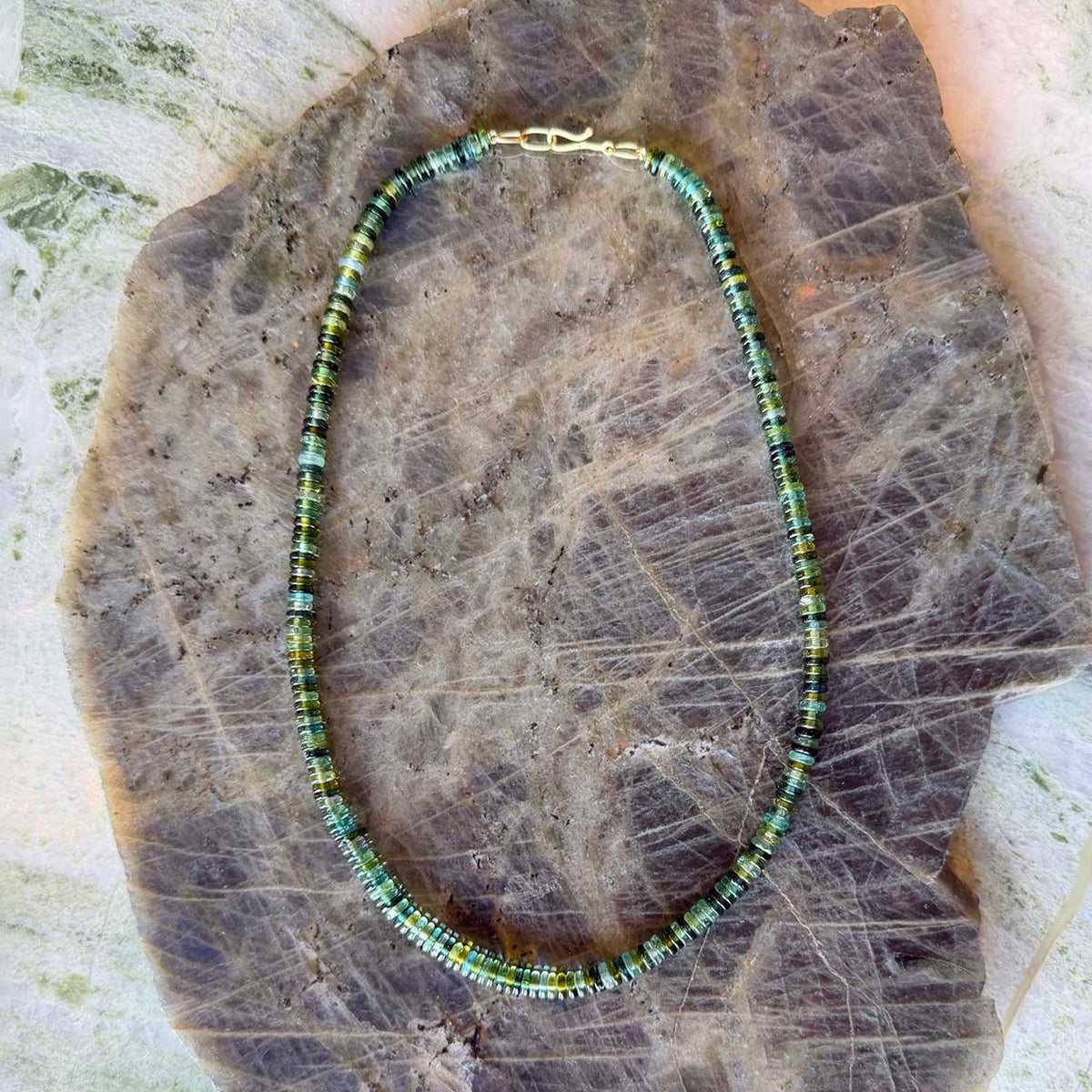 A beaded necklace made of small tourmaline beads in green, blue, and black, with a gold clasp, displayed on a textured brown stone slab. Made by Brooke gregson in Los Angeles.