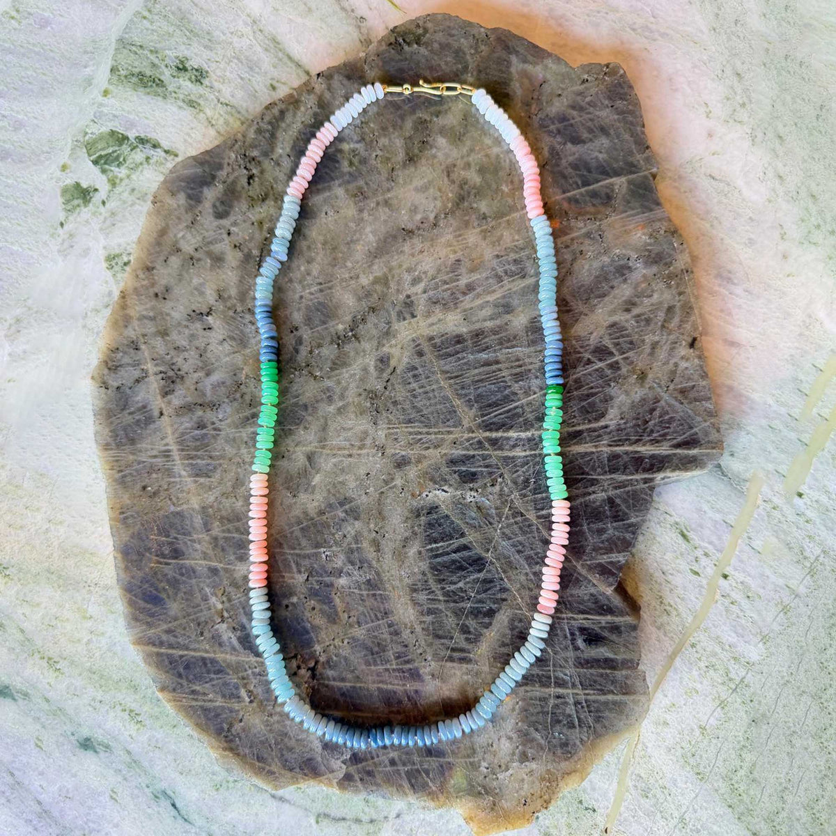 A beaded necklace made of small pastel beads in light blue, pink, green, and white, arranged in an oval on a textured brown stone slab.