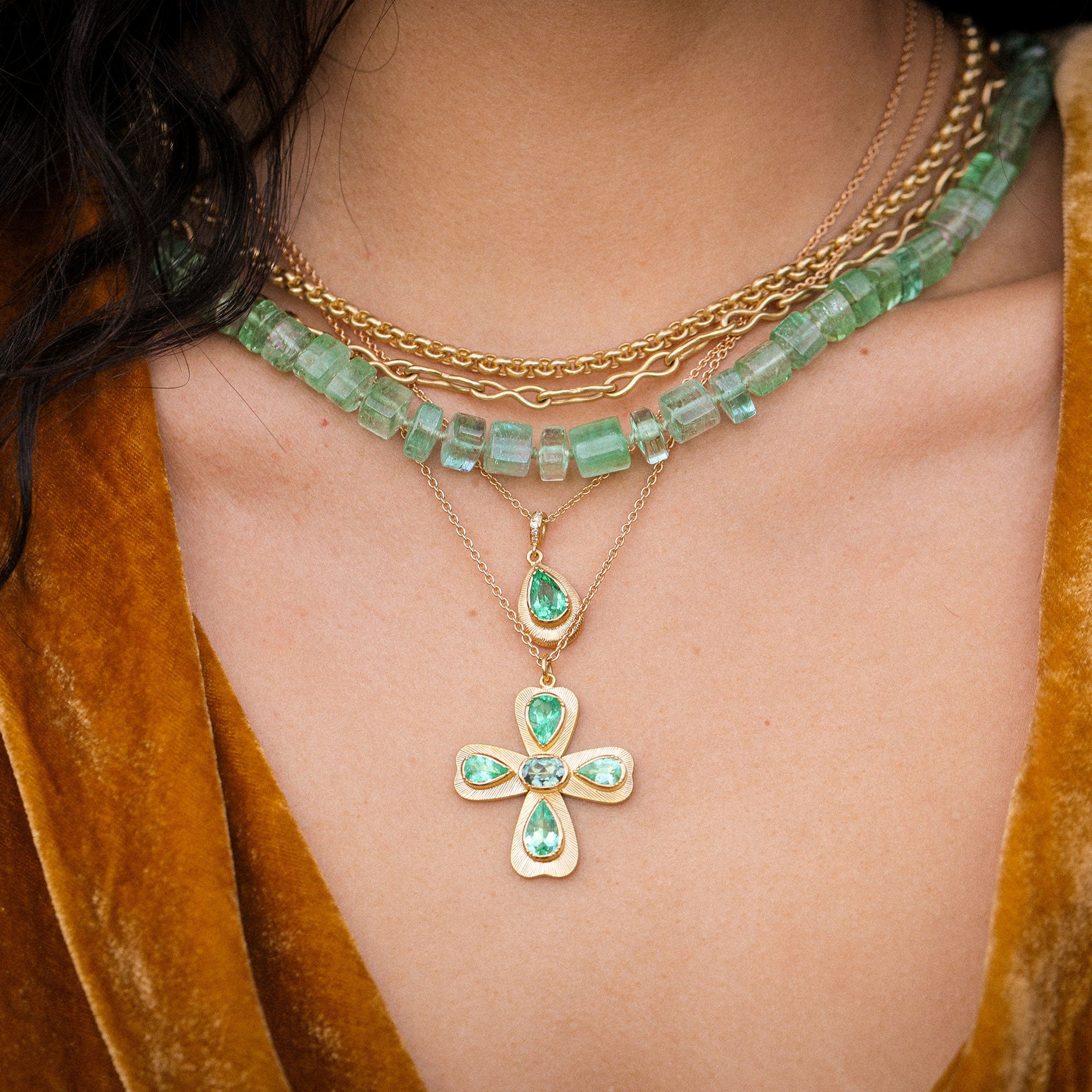 Necklace with green gemstones and gold cross pendant on a person wearing a brown garment.