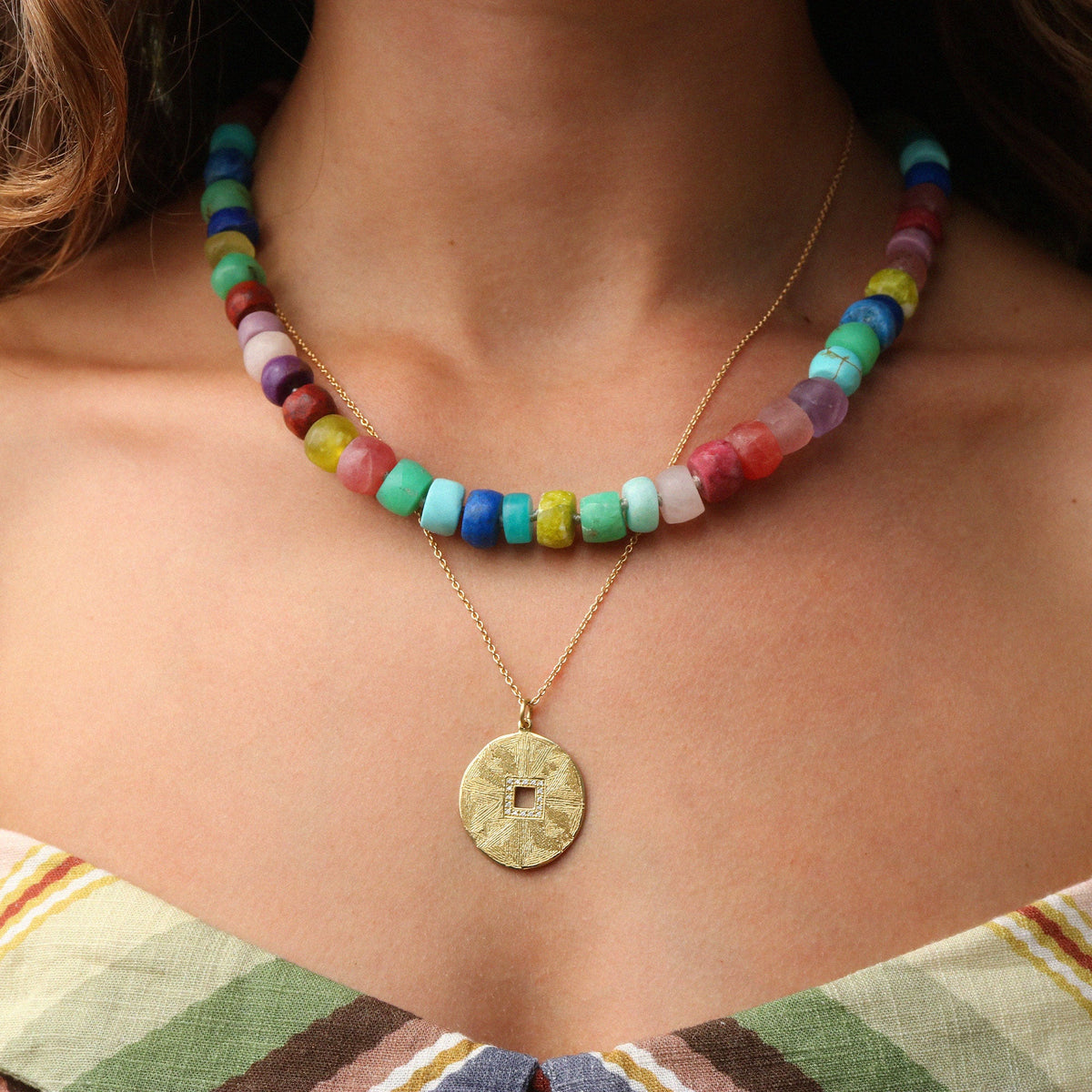 A close-up of a model wearing a multi-colored beaded necklace layered over a gold chain with a round textured coin pendant.