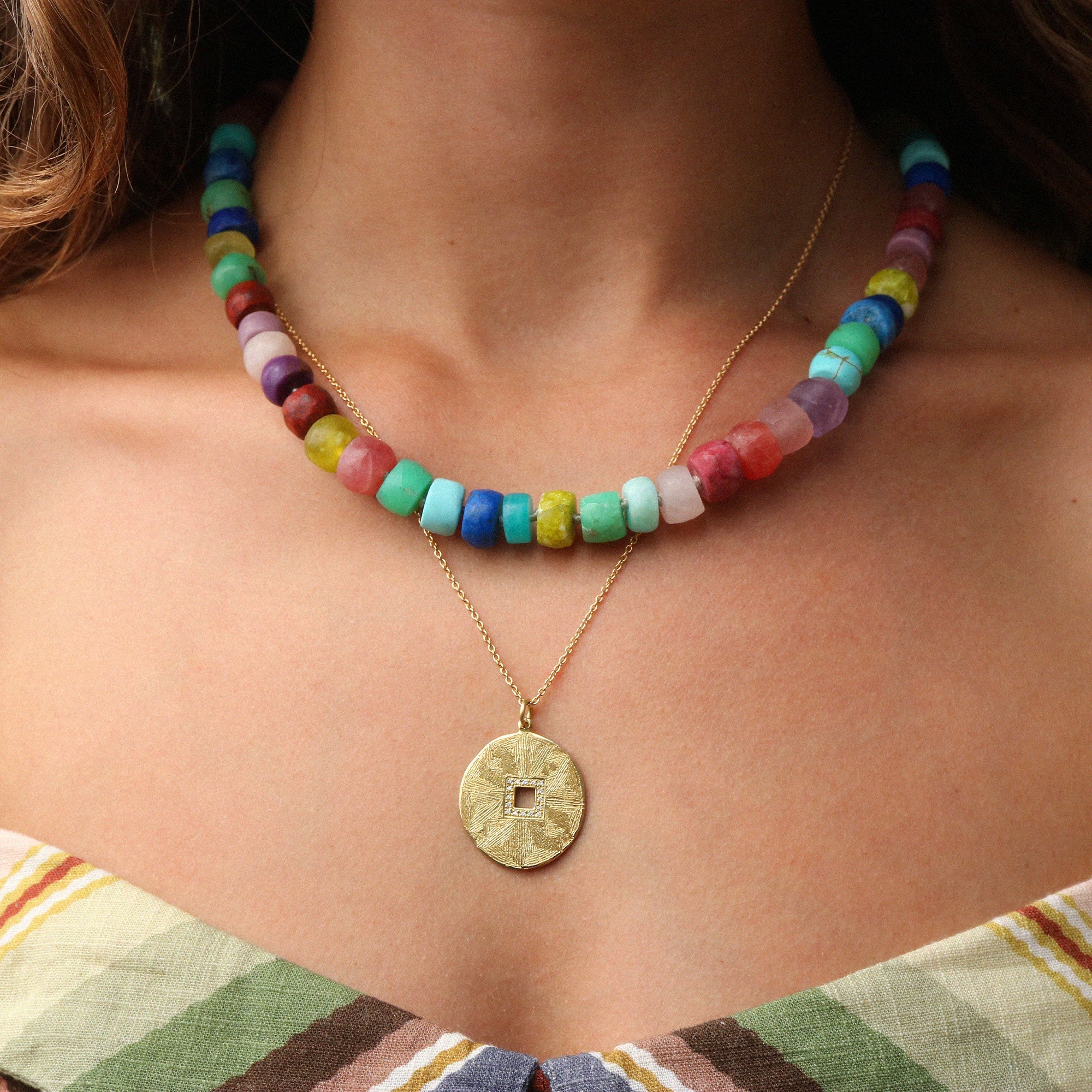A close-up of a model wearing a multi-colored beaded necklace layered over a gold chain with a round textured coin pendant.
