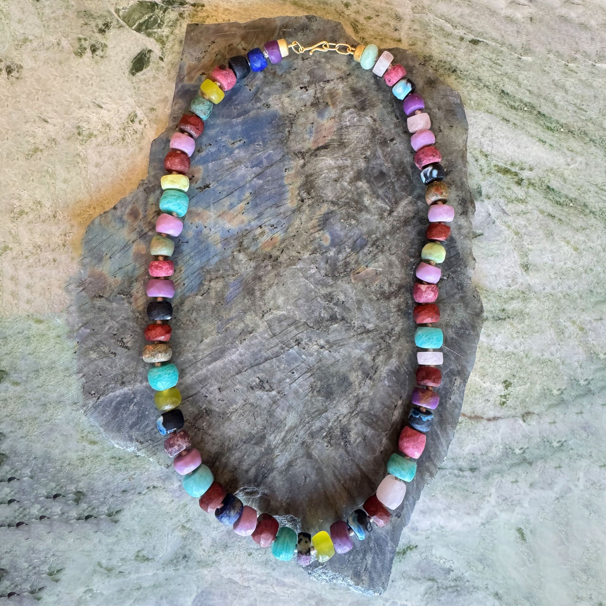 A beaded necklace made of many small, irregularly shaped polished beads in assorted colors, displayed on a large gray stone against a light stone surface.