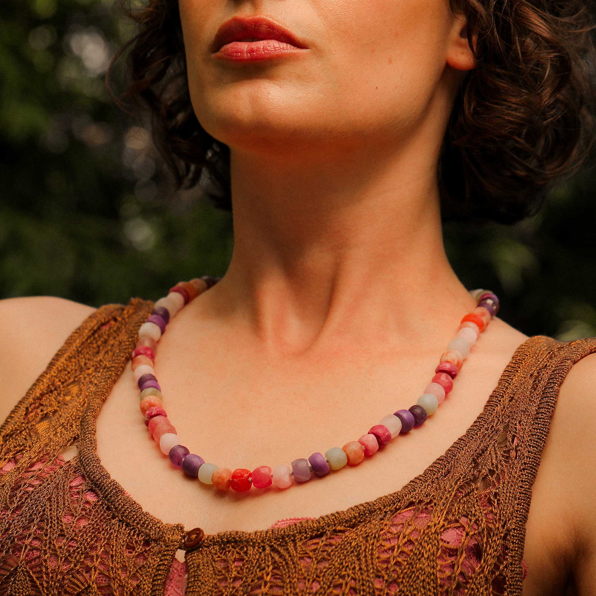 A close-up of a woman wearing a colorful beaded necklace with chunky round beads in shades of pink, purple, orange, and cream, paired with a brown crochet top against a blurred green outdoor background. Bead Necklace designed and made by Brooke Gregson in