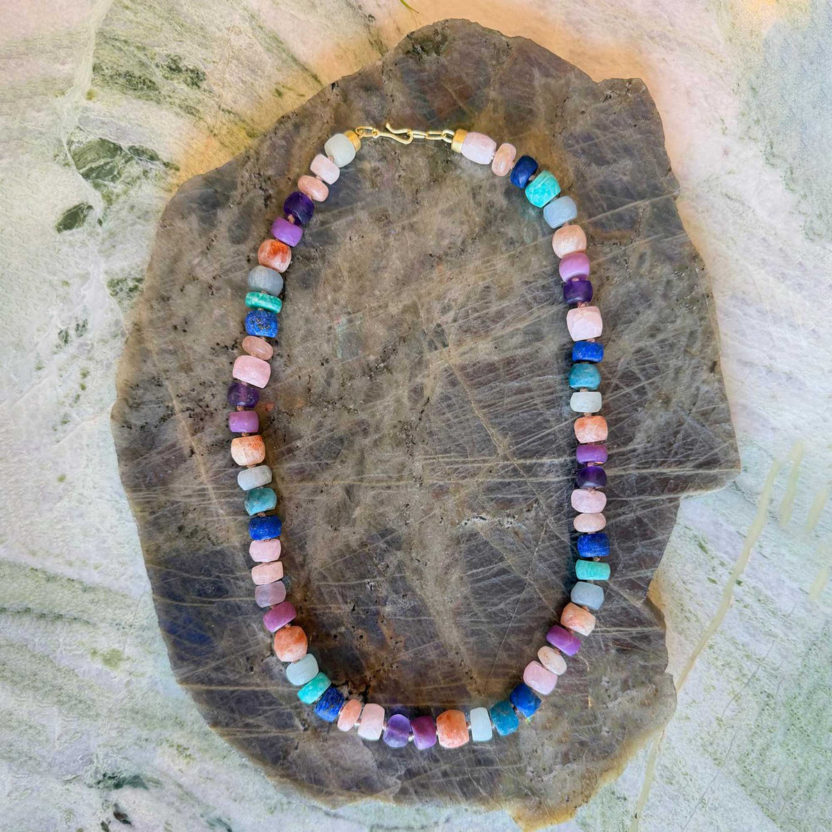 A beaded necklace made of small cylindrical gemstone beads in multiple colors including pink, blue, purple, green, and orange, displayed on a large polished brown stone against a light textured background.