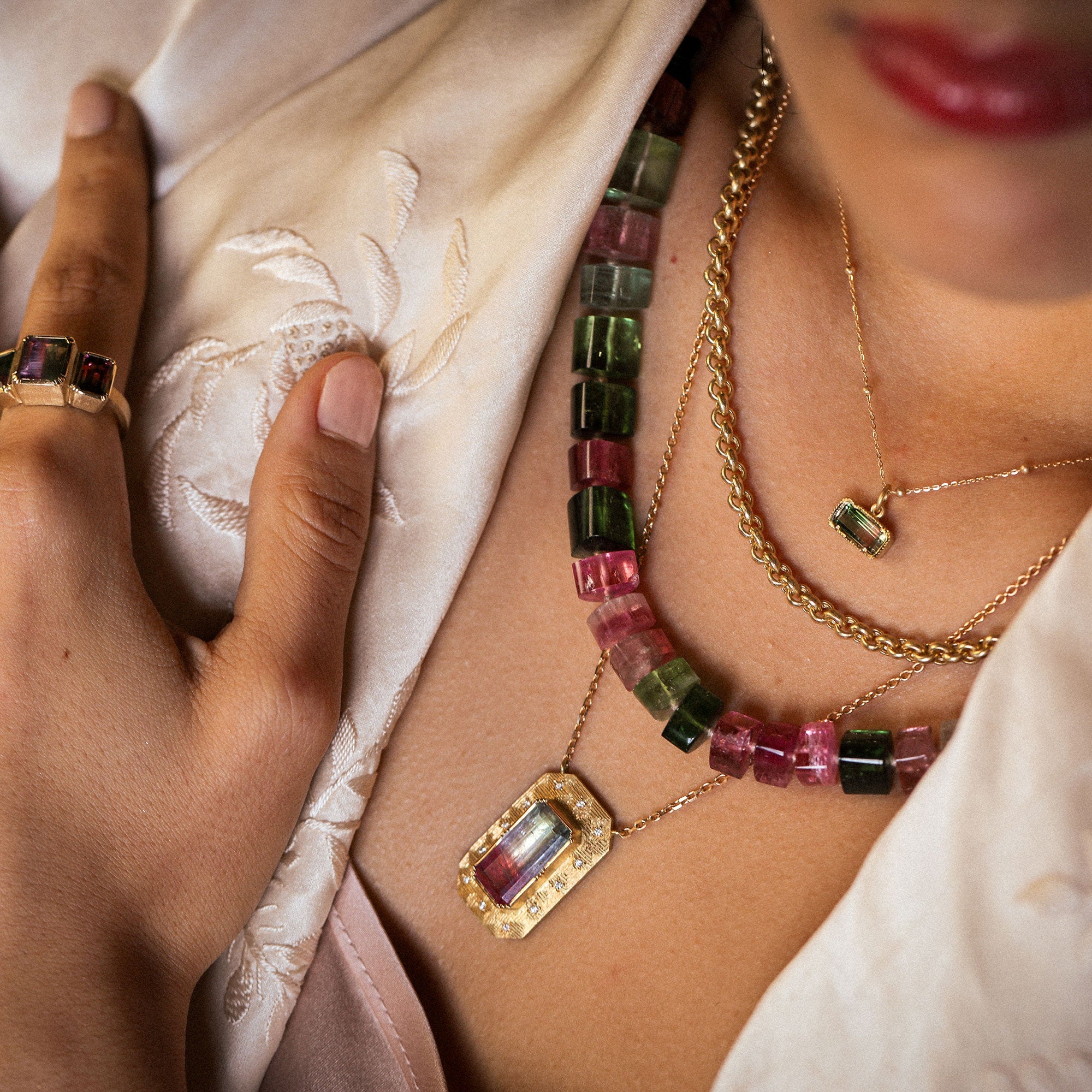 Close-up of a person wearing a colorful gemstone necklace and gold chain, with a hand touching a white fabric background.