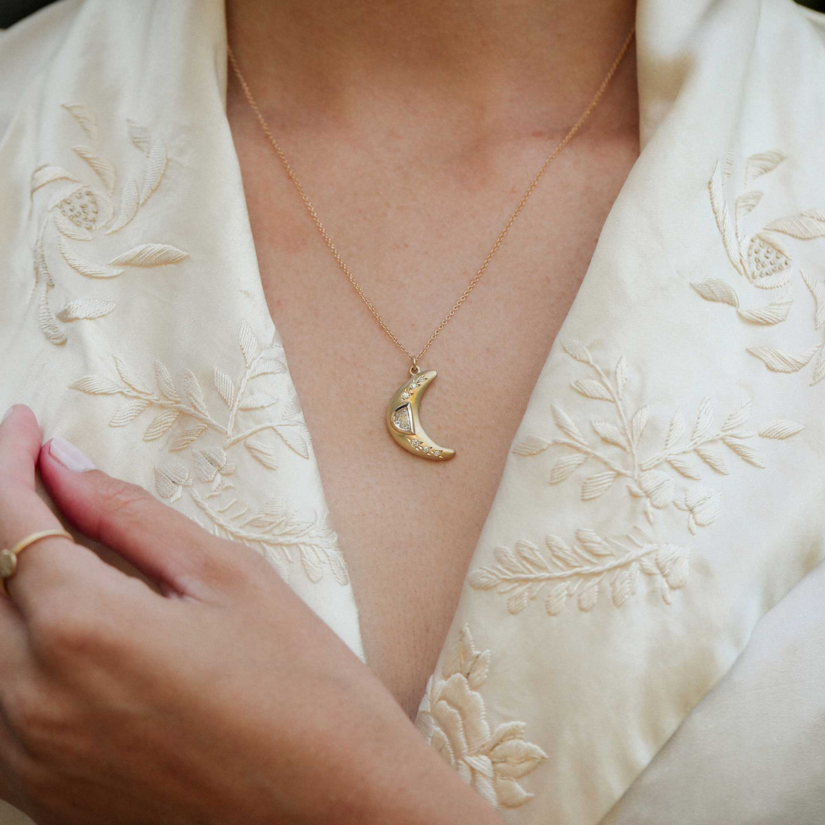 A close-up of a person wearing a delicate gold crescent moon pendant necklace with white diamonds, styled with a cream-colored embroidered satin robe. designed by brooke gregson in los angeles.