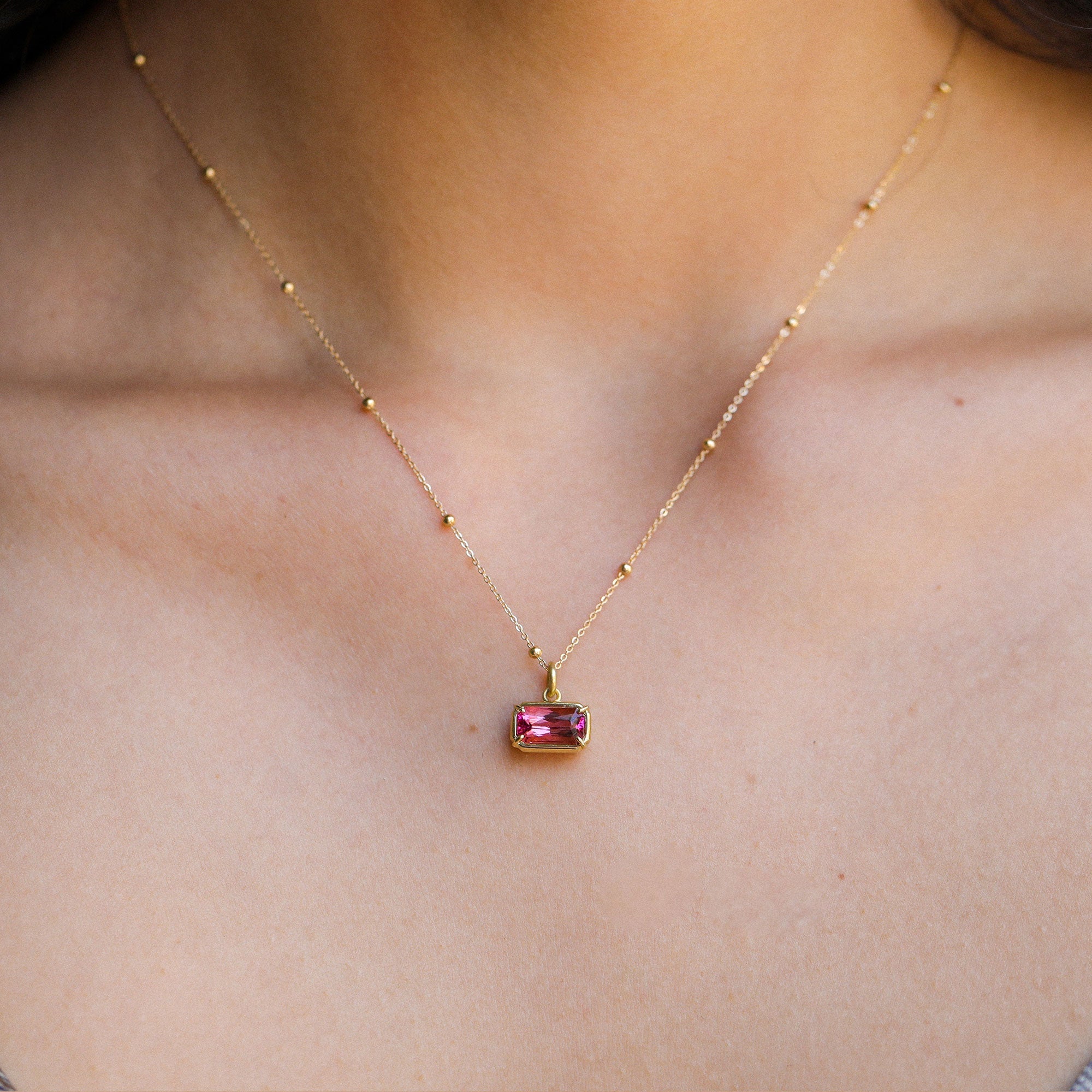 A delicate gold chain necklace with a small rectangular pink tourmaline gemstone pendant, shown worn on a person's neck and upper chest.