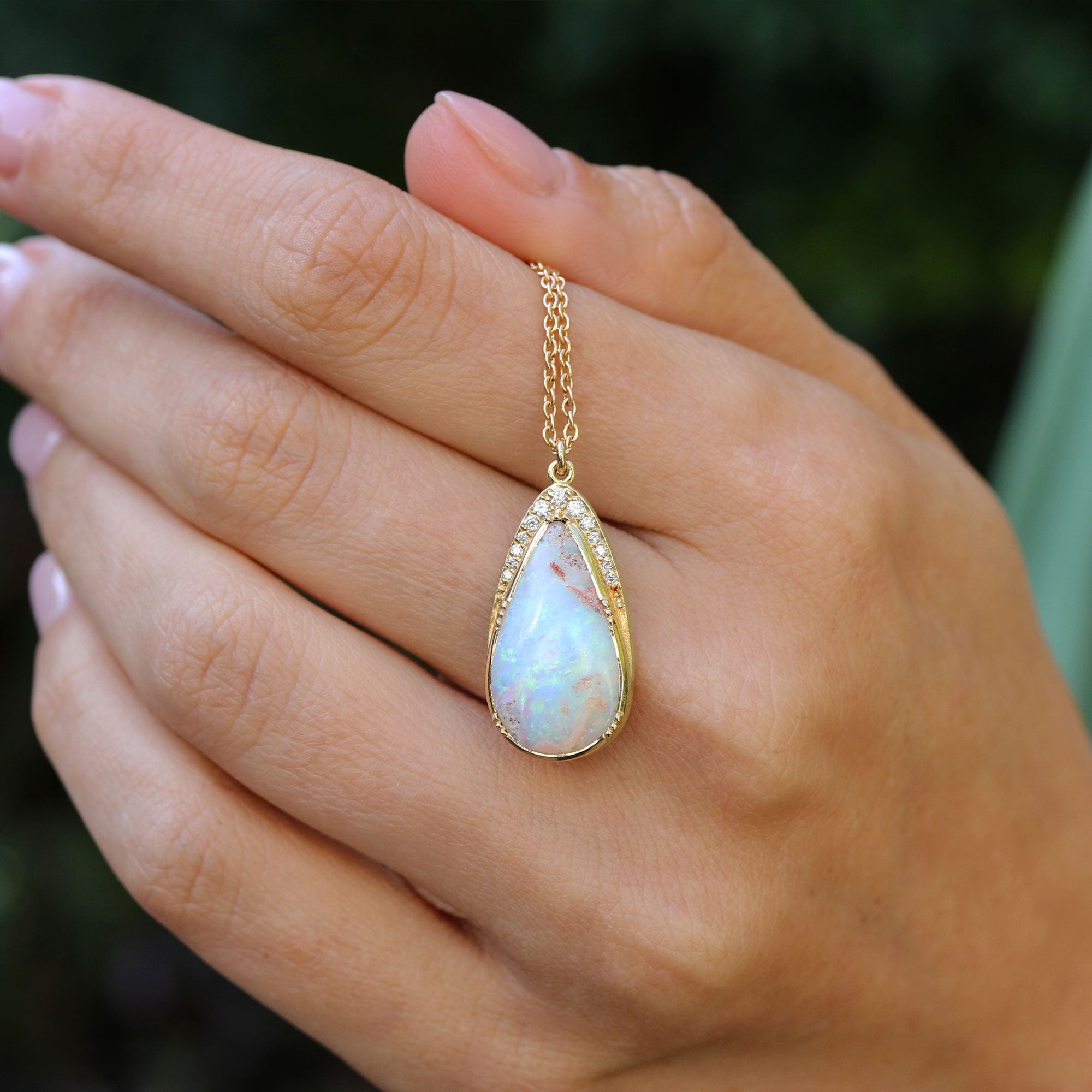 Hand holding a gold necklace with a teardrop-shaped opal pendant against a blurred green background