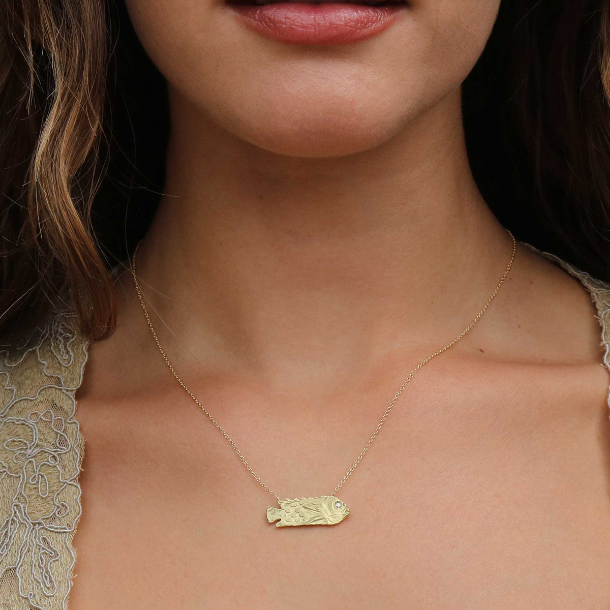 A close-up of a model wearing a delicate gold 18k chain necklace with a textured, fish-shaped pendant by brooke gregson jewelry.