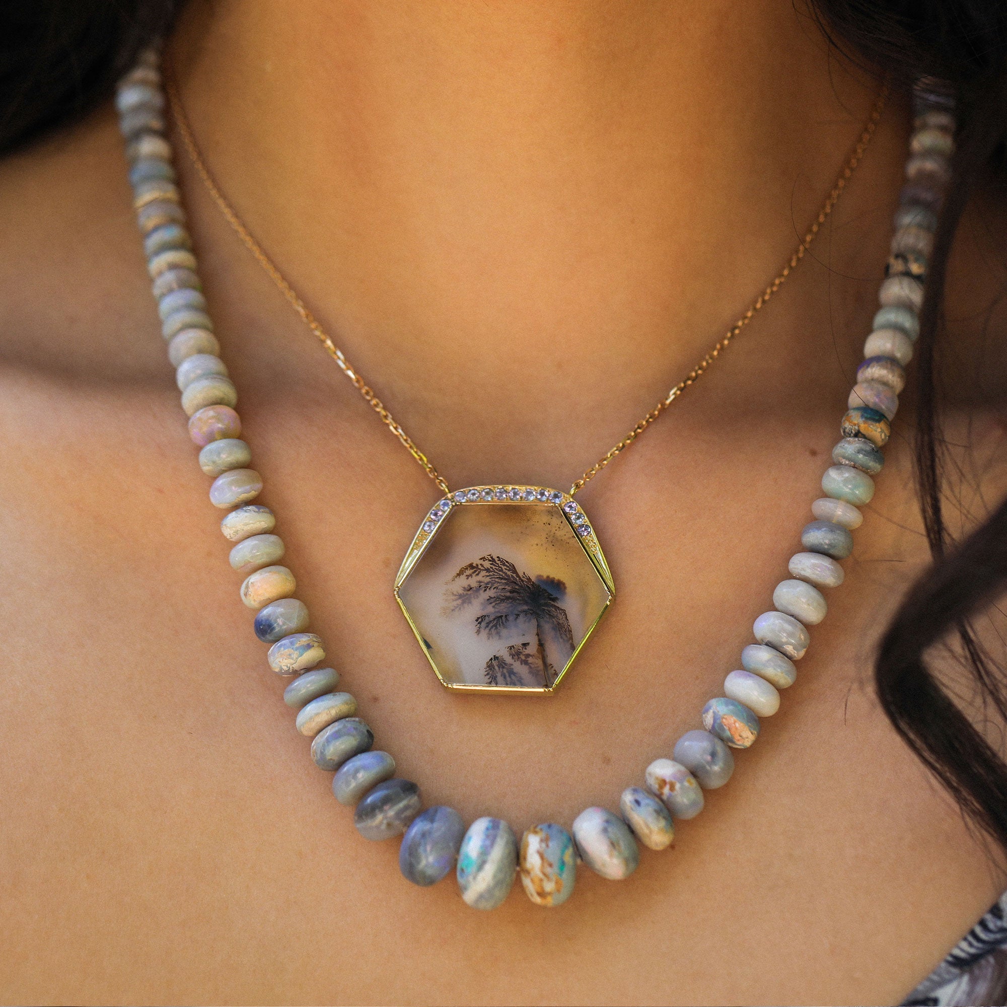 A close-up of a person's neck and chest wearing a layered necklace. The necklace features a gold chain with a hexagonal dendritic agate pendant containing a small scenic photograph, layered with a strand of round, marbled blue-gray beads.