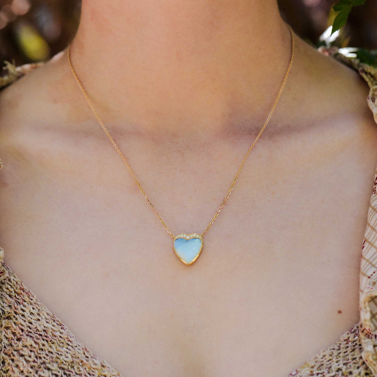 A gold chain necklace with a small heart-shaped pendant featuring a pale blue boulder opal inlay with white diamond accents, worn on a model's neck.