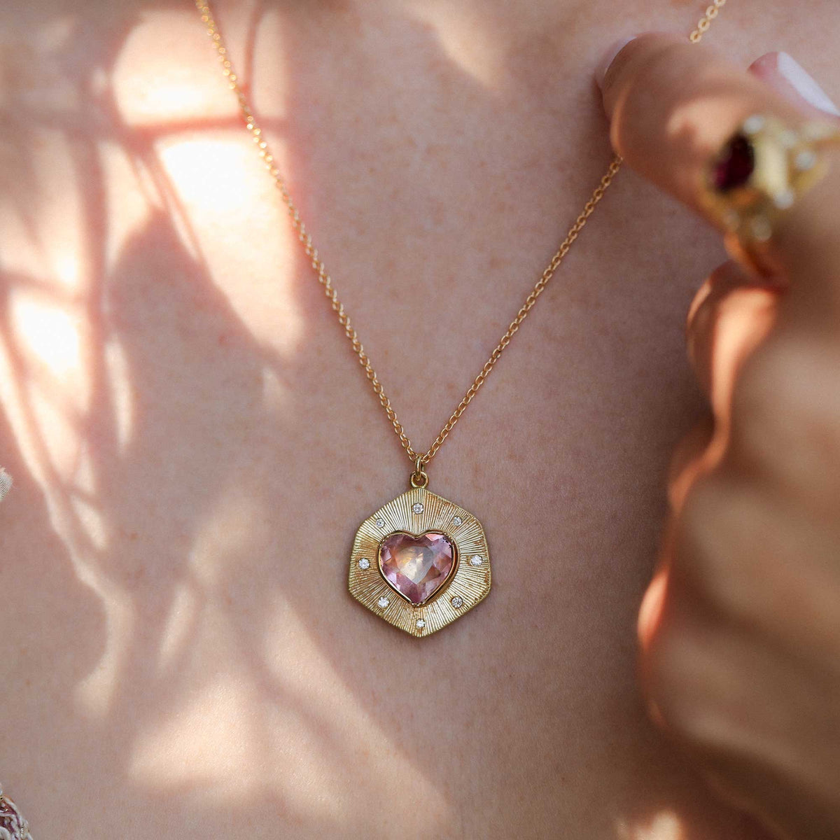 A gold necklace with a textured hand engraved hexagonal pendant featuring a pink heart-shaped tourmaline gemstone and small diamond stones, worn on a person's neck with warm sunlight and leaf shadows.