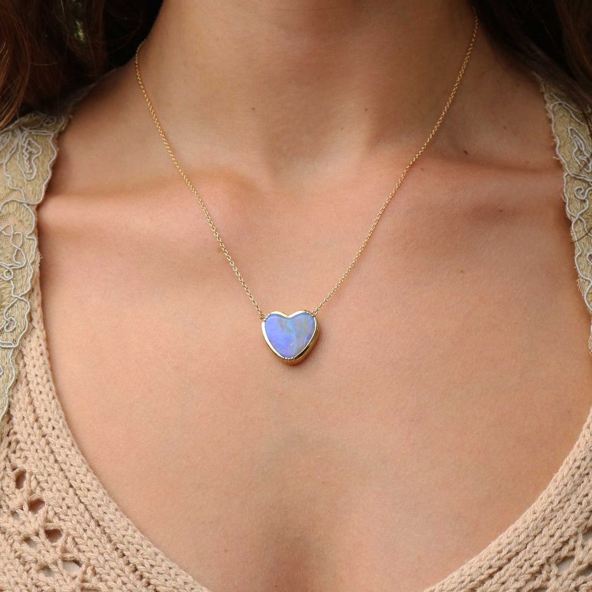 A gold heart-shaped boulder opal pendant necklace with a light blue iridescent opal center, worn on a model's neck over a beige lace-trim top.