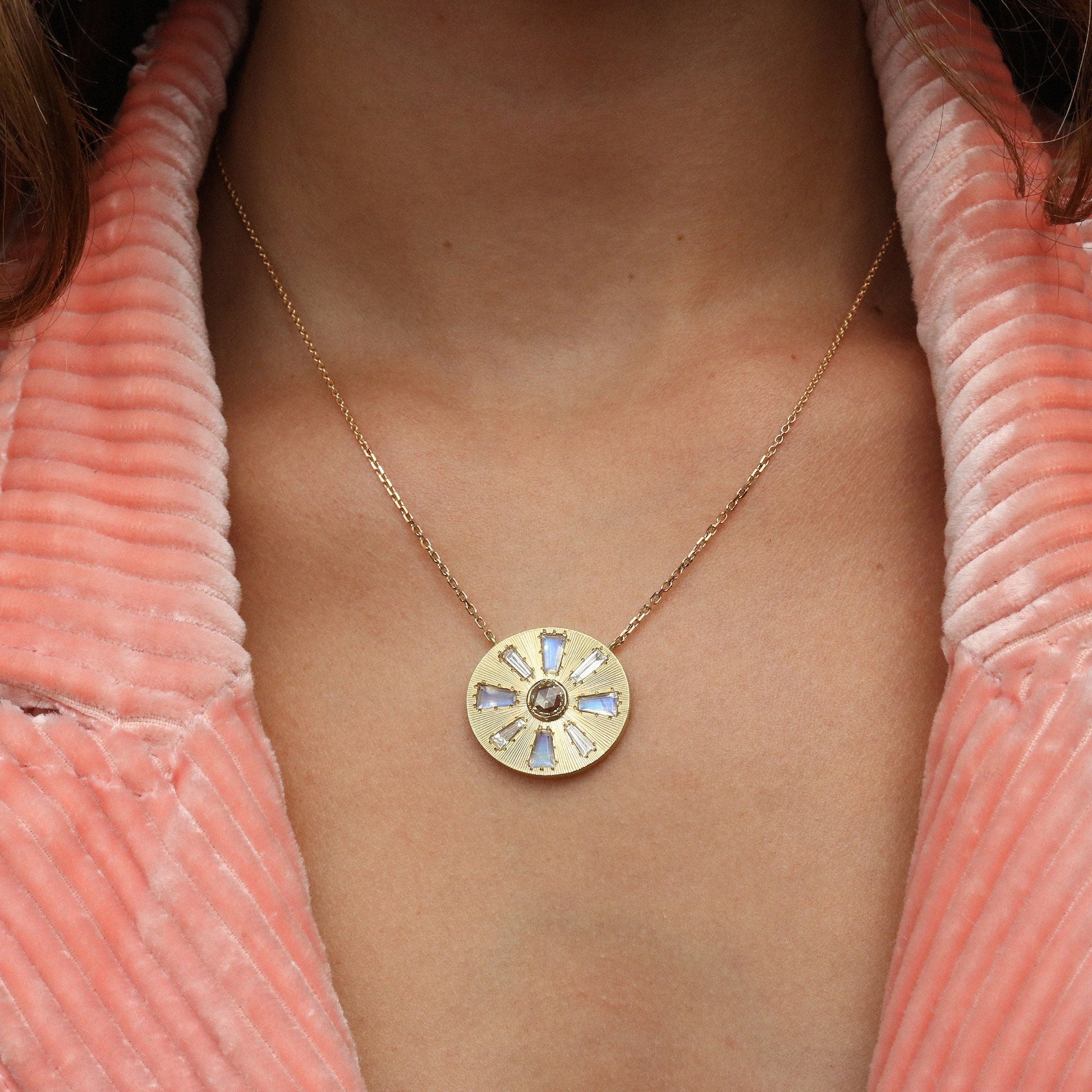 A gold circular pendant necklace with a sunburst diamond design and moonstone inlay, worn on a model's neck over a pink ribbed top.
