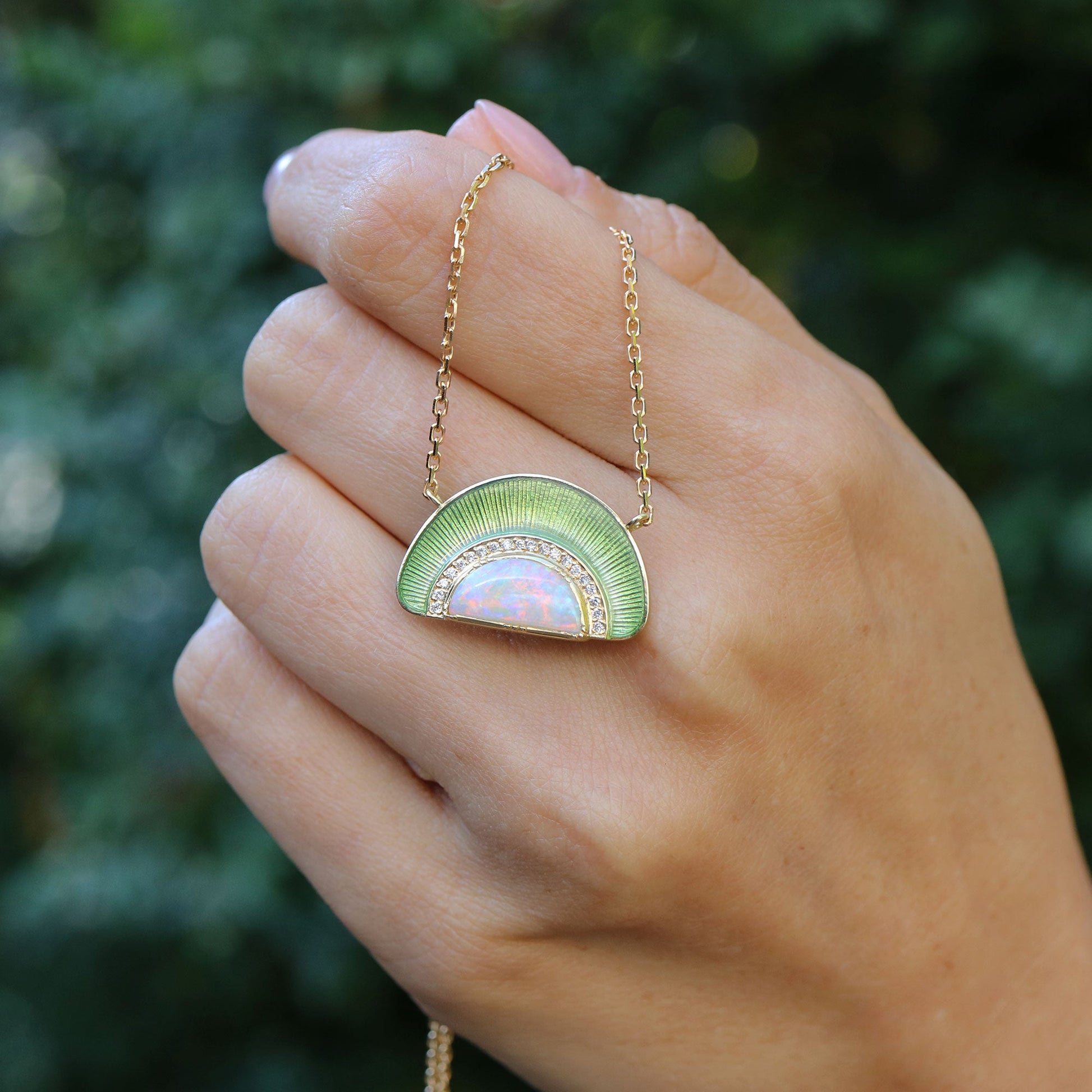 Hand holding a 18k gold necklace with a pendant with opal, white diamonds and sage green enameling by Brooke Gregson.
