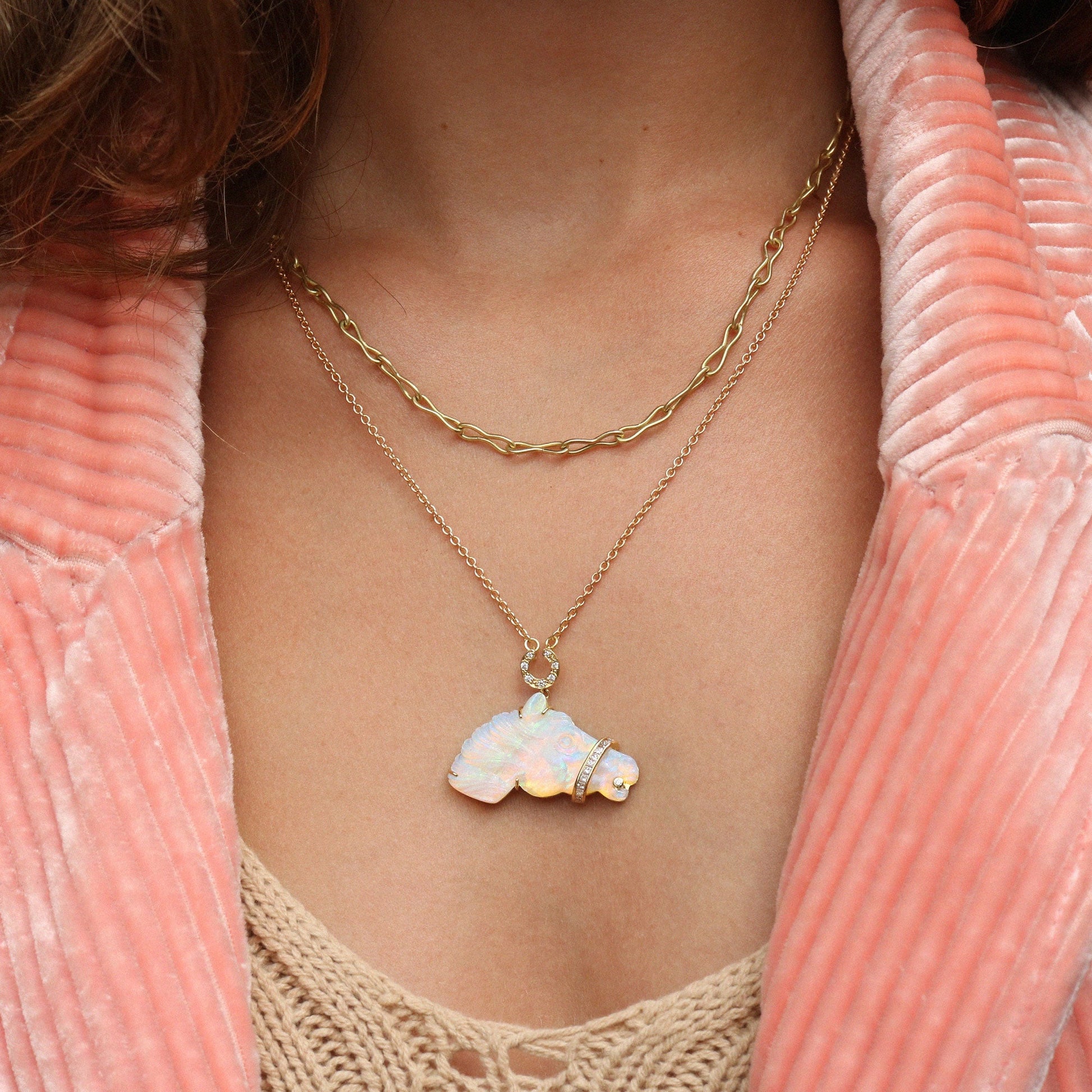 A close-up of a person wearing a layered necklace set. The top layer features a gold chain with a small round clear stone pendant, and the bottom layer features a gold chain with a white iridescent opal horse pendant.