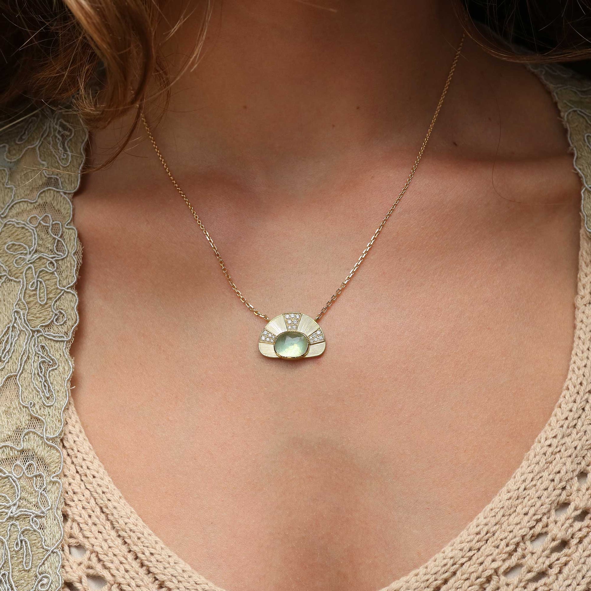 A gold necklace with a semi-circular pendant featuring a light green aquamarine center stone and small diamond stones along the top curve, worn by a model in a beige lace and knit top.