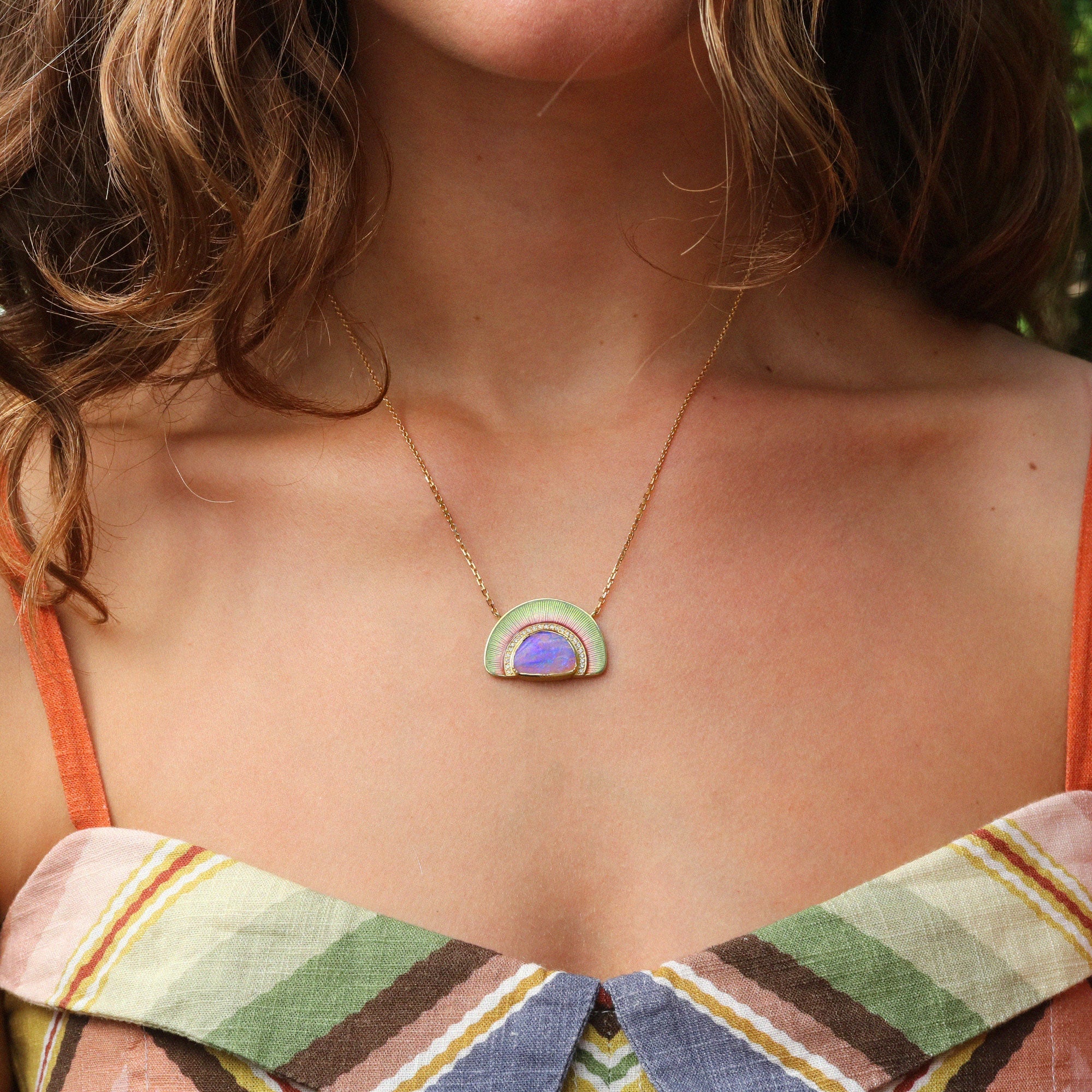 A close-up of a woman wearing a gold chain necklace with a small arch-shaped pendant featuring light green, lavender purple, and pink enamel details.