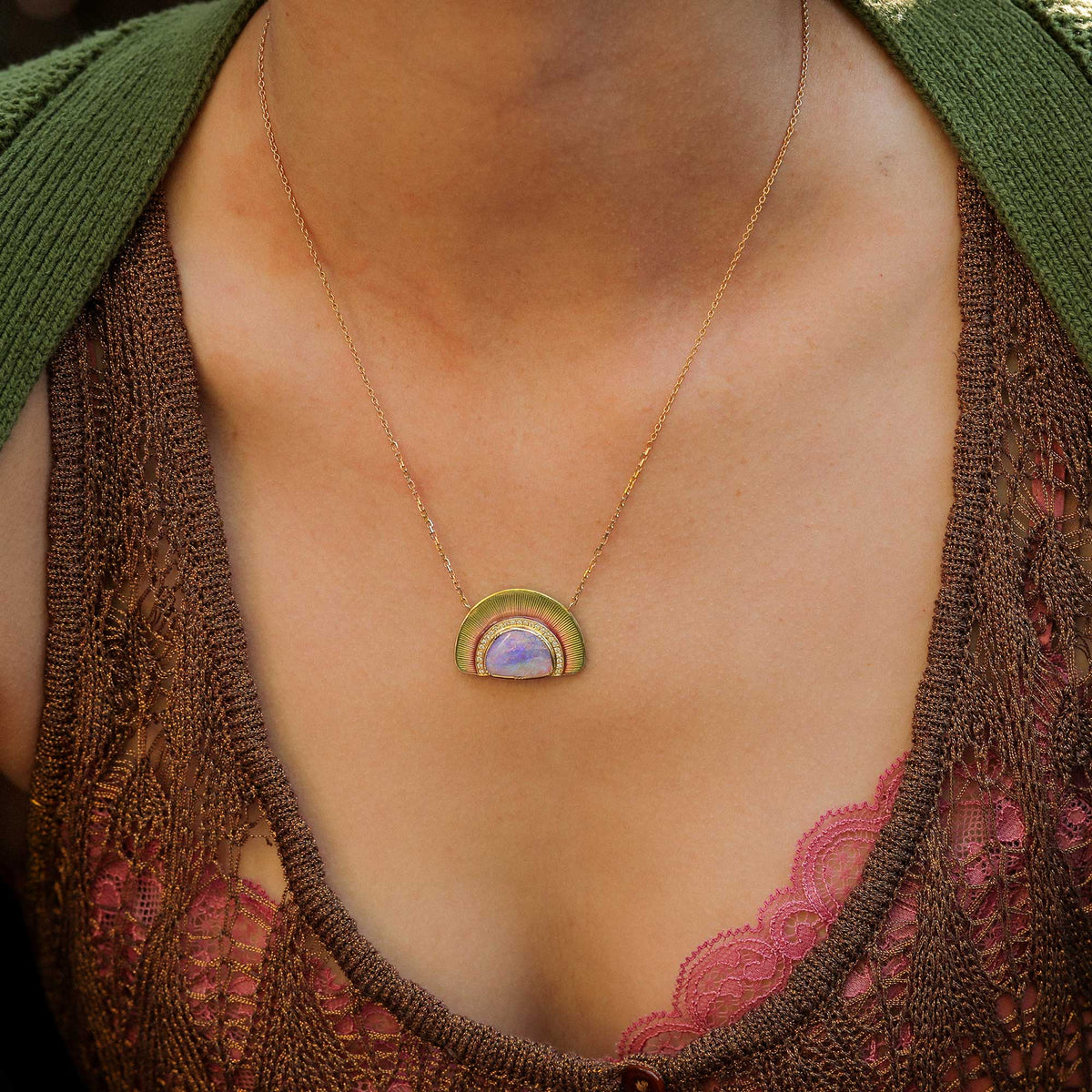 A close-up of a person wearing a delicate gold chain necklace with a small rainbow enamel pendant. The pendant features concentric arches in olive green, gold, and pink, with a smooth lavender-purple opal center with white diamond pave. designed in london