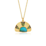 An 18k yellow gold pendant necklace featuring a fan-shaped design with a central iridescent blue-green opal and small emerald and sapphire gemstone accents with hand engraving, shown on a white background with a soft reflection. By jewelry designer Brooke