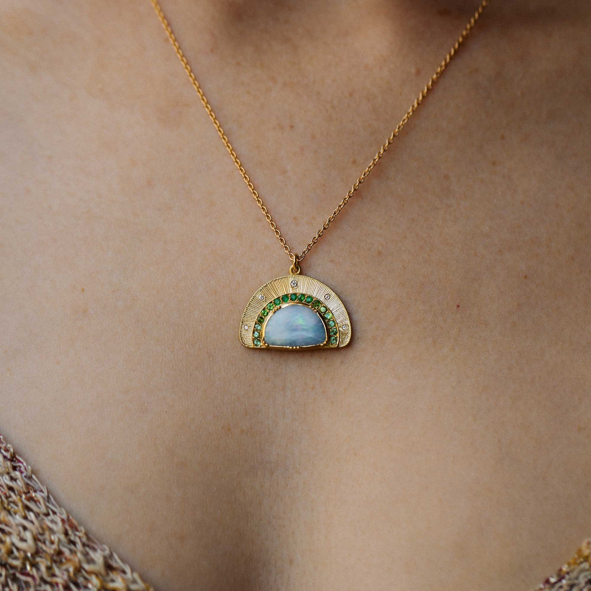 A gold 18k necklace with a semi-circular pendant featuring a light blue opal center stone and green emerald and tsavorite stone accents, worn on a model's neck. designed by brooke gregson.