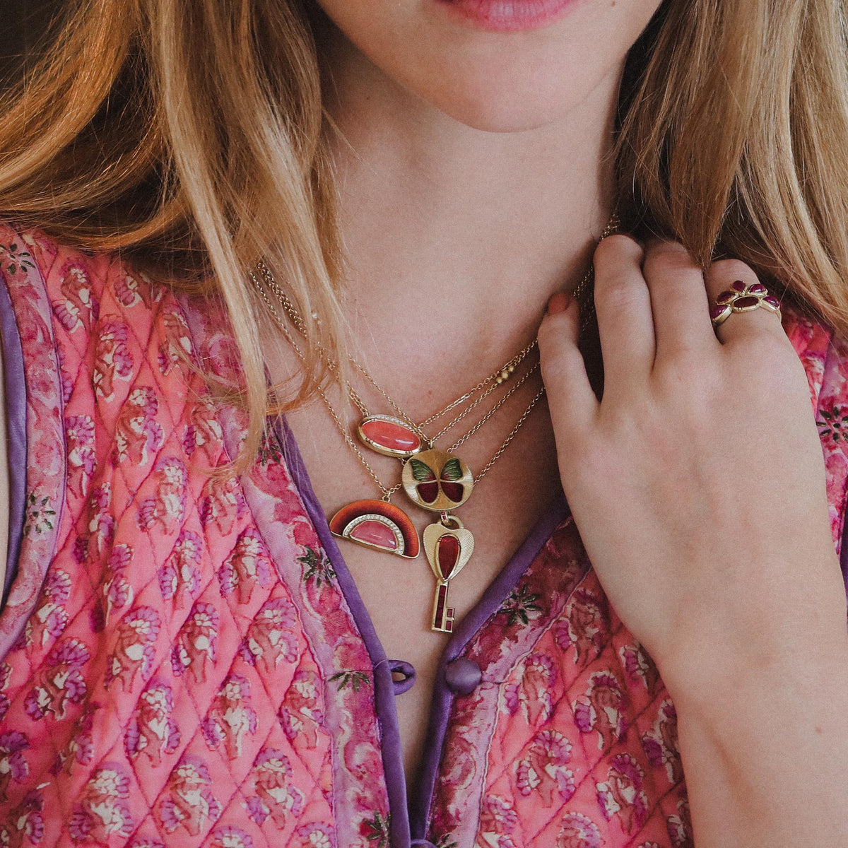 A close-up lifestyle shot of a woman wearing a layered gold necklace set with colorful enamel charms, including a rainbow, a key, and a clover motif, paired with a pink floral quilted top. Jewelry by Brooke Gregson.