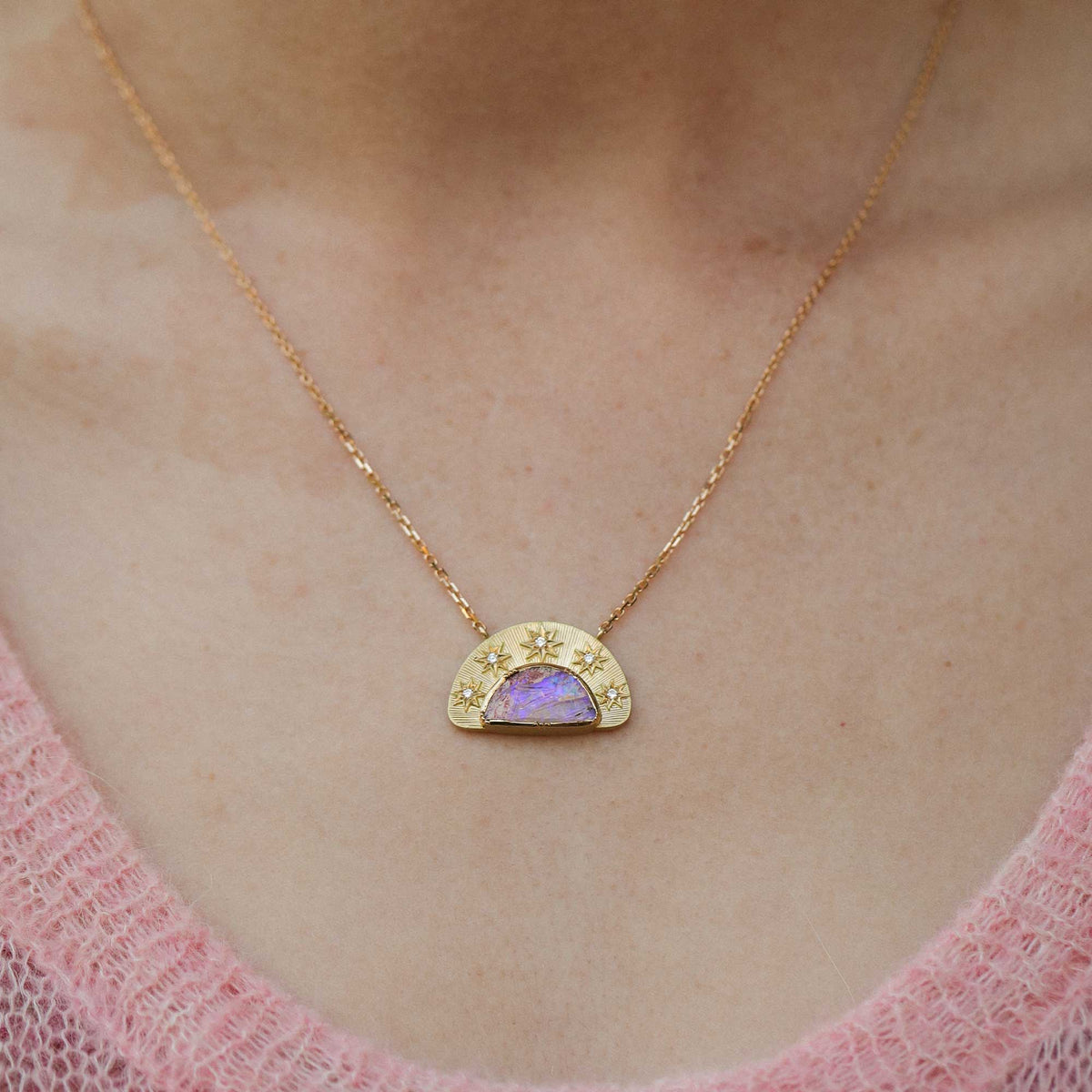 A gold necklace with a crescent moon pendant featuring a purple iridescent opal center stone and small star diamond details, worn on a person's neck over a pink knit top.