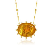 A gold pendant necklace featuring a large oval citrine gemstone with a sculpted sun face in the center, surrounded by small clear stones, on a gold beaded chain.