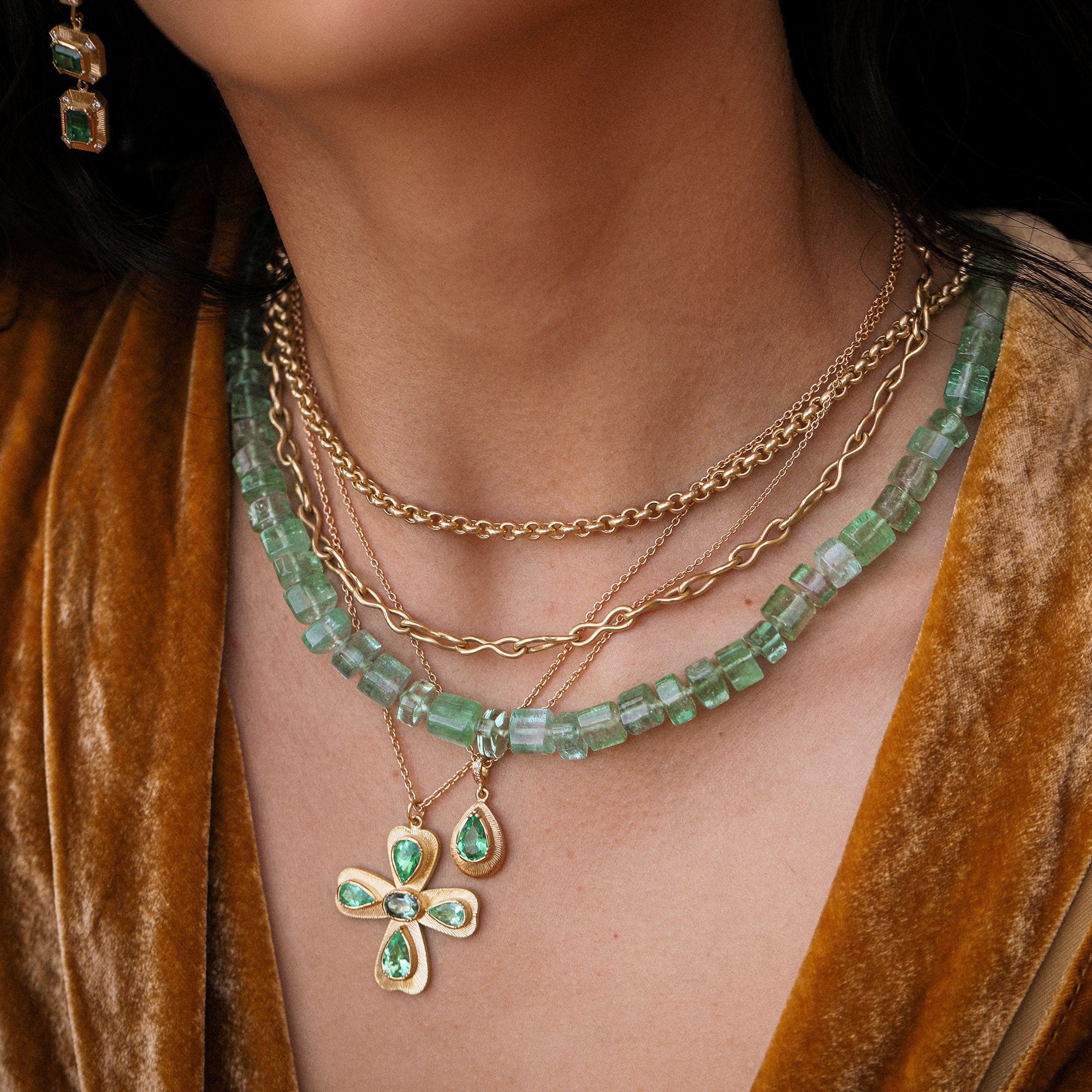 Close-up of a person wearing a gold necklace with green gemstones and a cross pendant, against a brown velvet background.