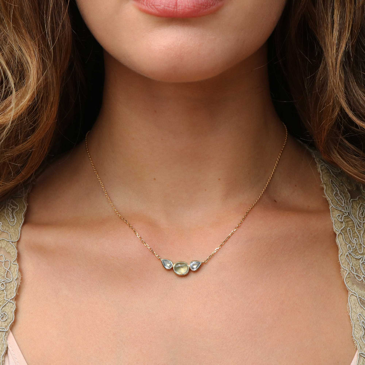 A close-up of a model wearing a delicate gold chain necklace with three small teardrop-shaped gemstones in a row, set against her neck and upper chest.