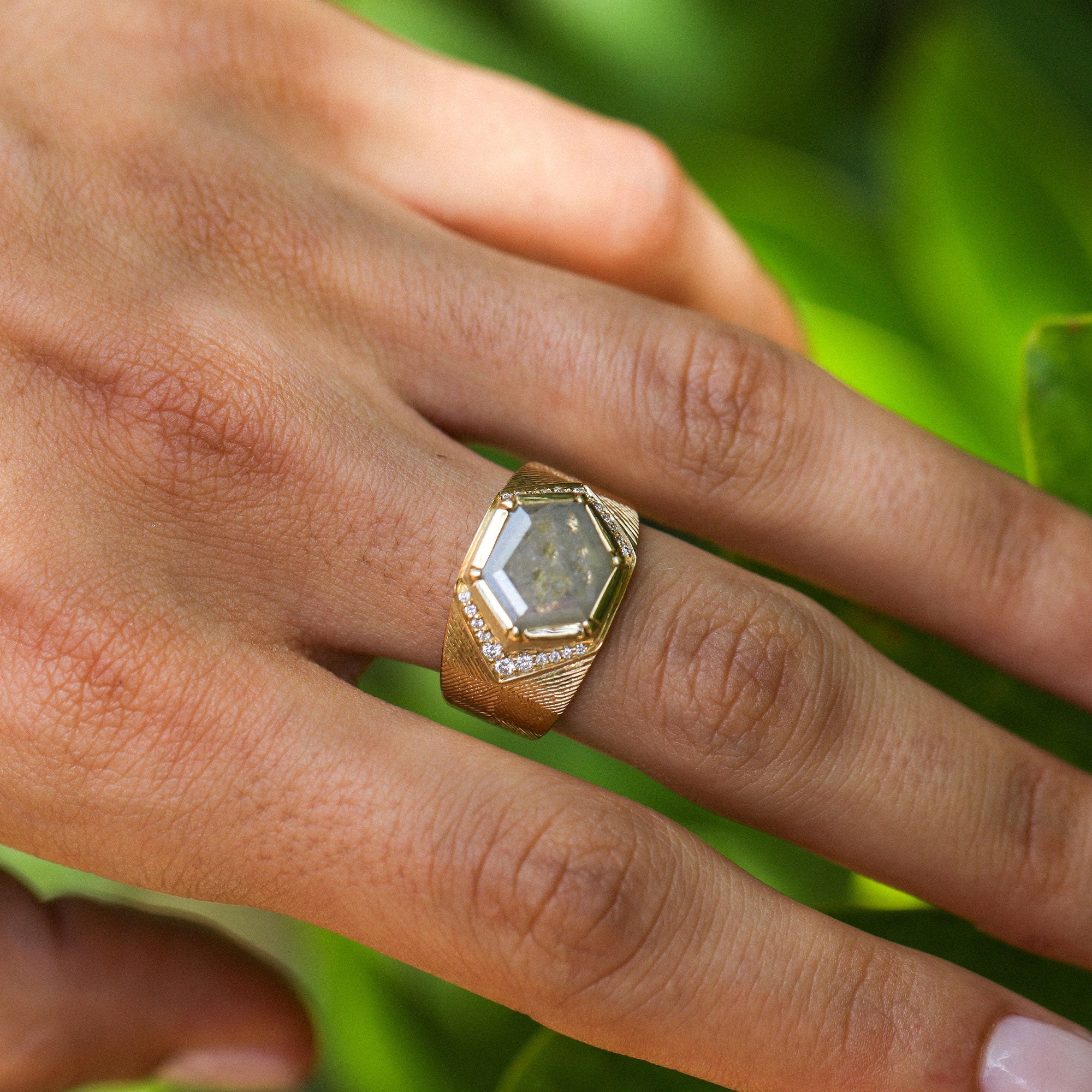 A gold ring with a faceted clear raw diamond gemstone and small pavé diamond accents, shown worn on a person's finger against a blurred green background.