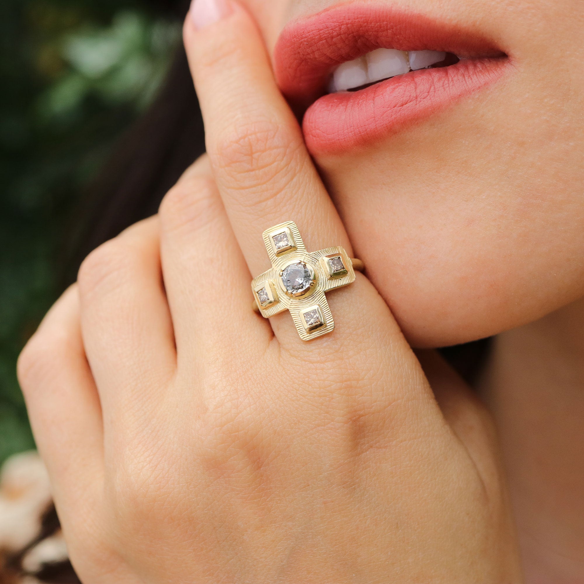 Close-up of a hand wearing a gold ring with diamond gemstones, against a blurred natural background.