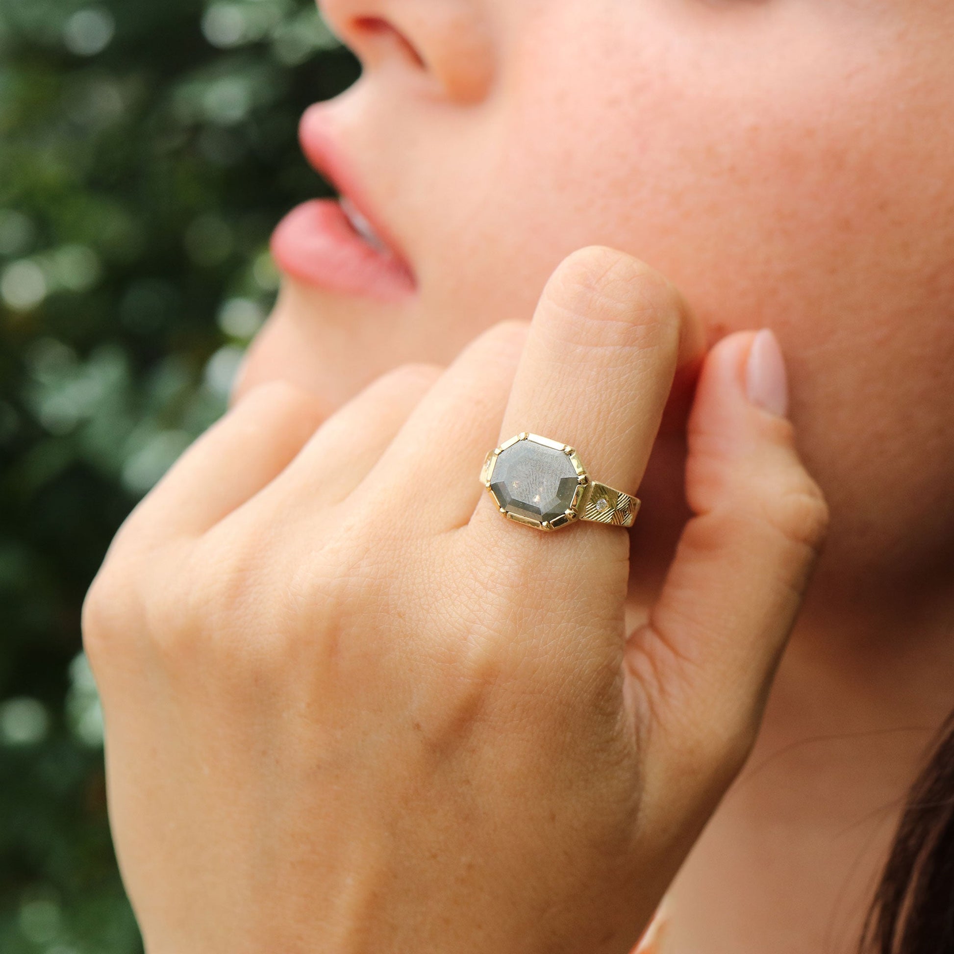 Close-up of a hand wearing a gold ring with a large raw diamond gemstone, touching the cheek against a blurred green background.
